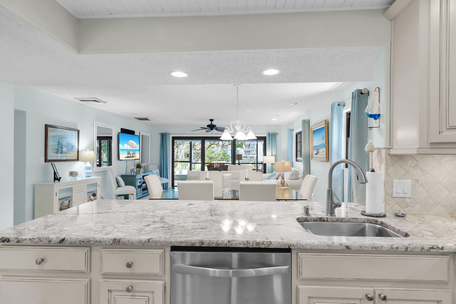Open-plan kitchen and living area with a marble island, stainless dishwasher, and large curtained windows.
