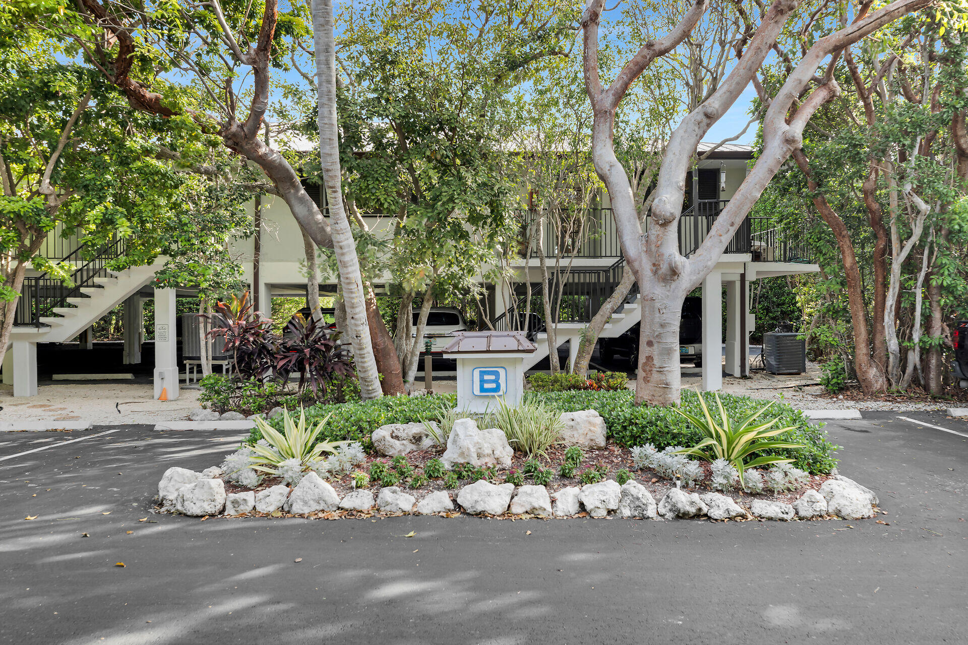 Raised residential building partly screened by tropical trees, with exterior stairs, balconies, and a landscaped entrance.