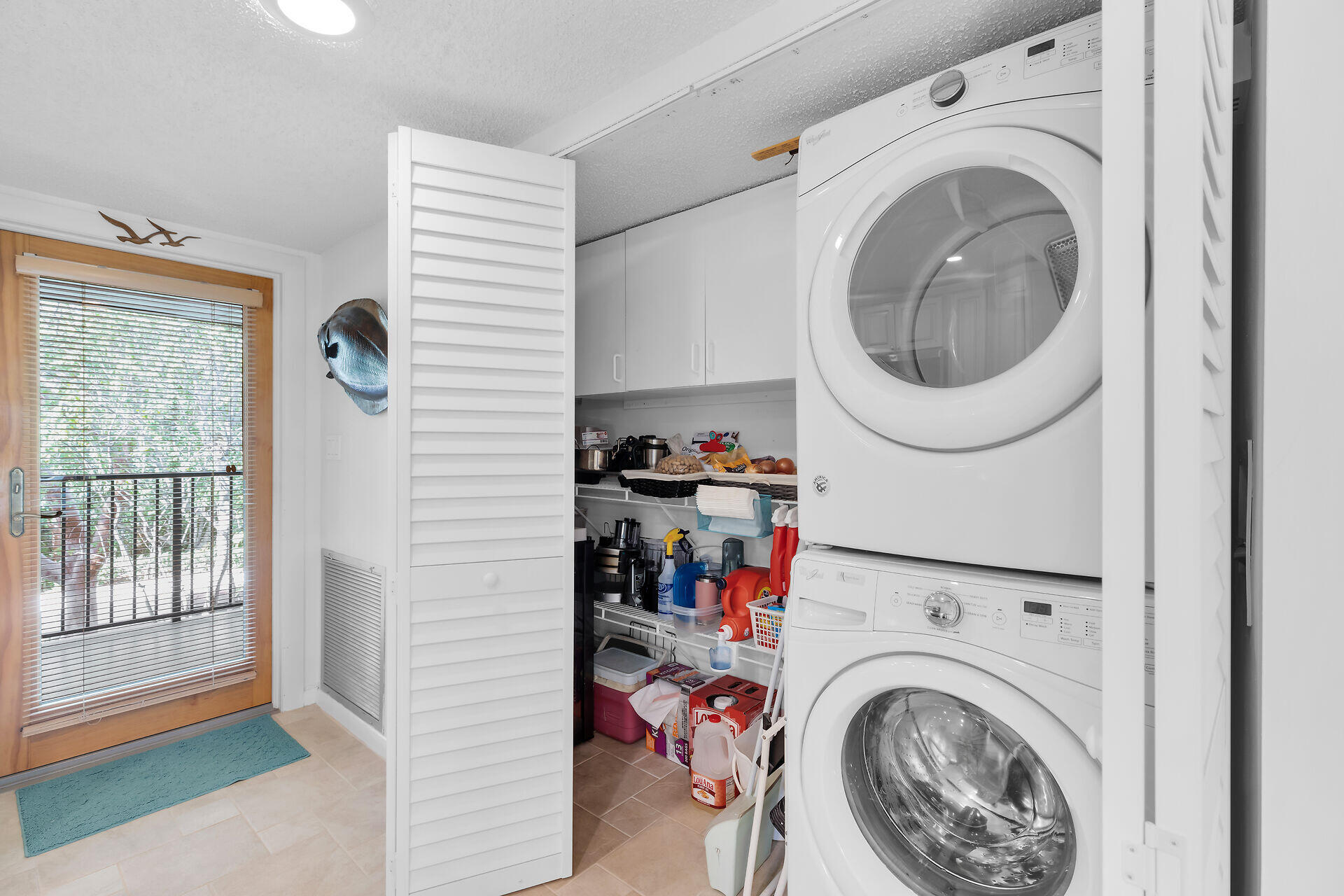 Laundry area with stacked washer and dryer, open storage closet, and wood-framed glass door with blinds.