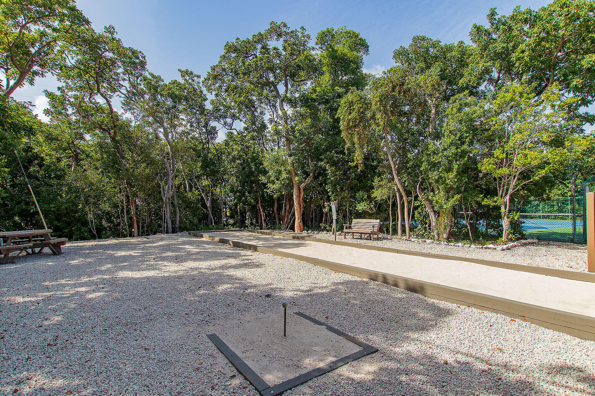 Gravel bocce court with benches and a picnic table beneath trees, with a fenced tennis court nearby.