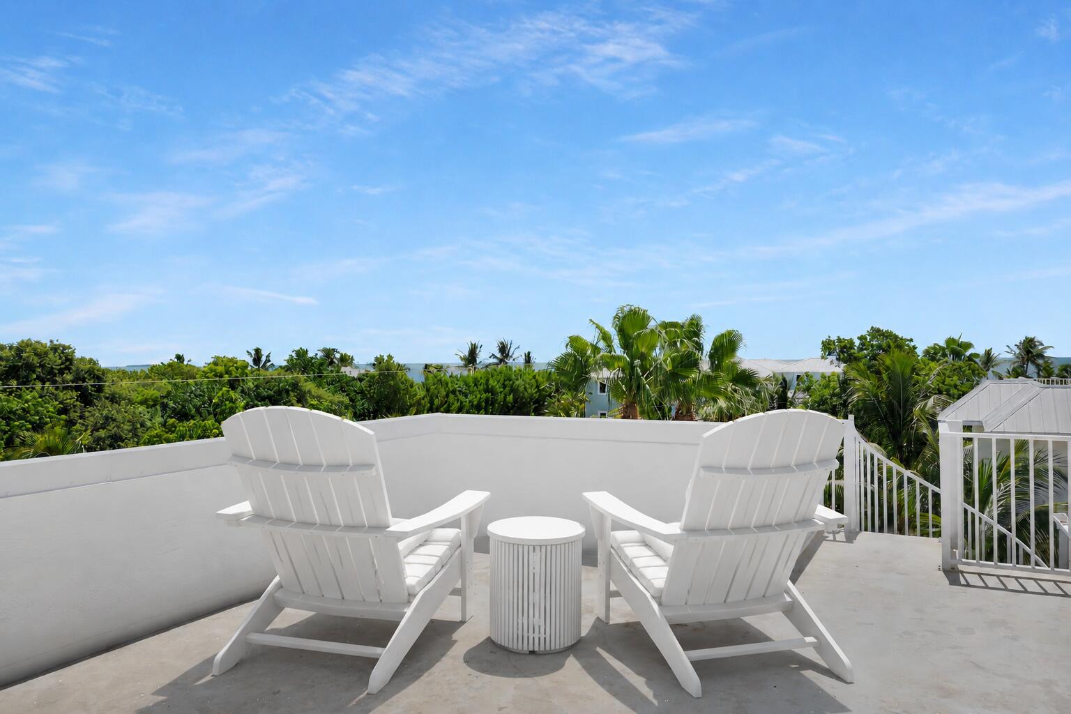 Rooftop terrace with two white Adirondack chairs and a round table overlooking tropical greenery.