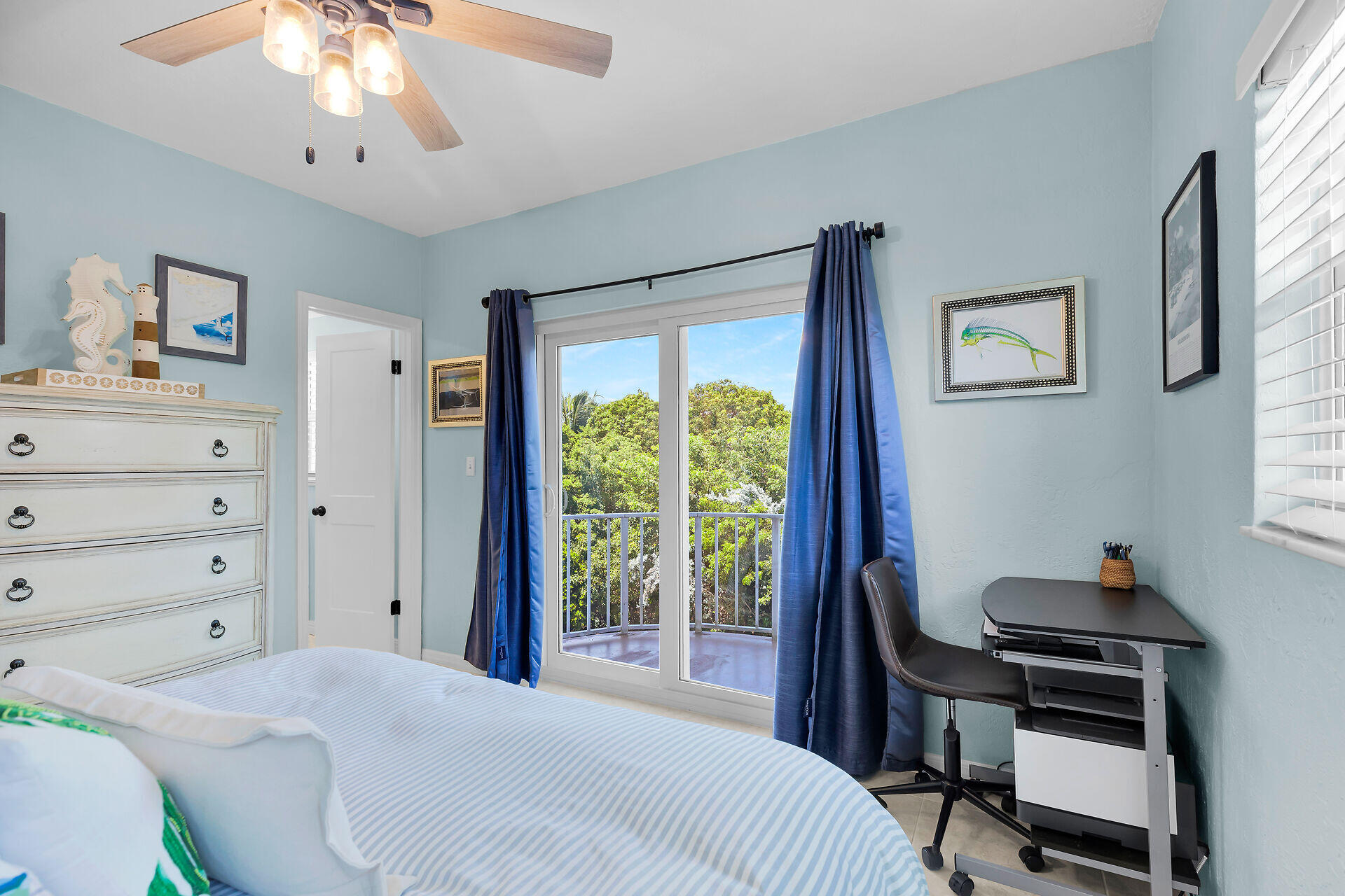 Bedroom with blue curtains framing sliding doors to a balcony overlooking leafy trees.