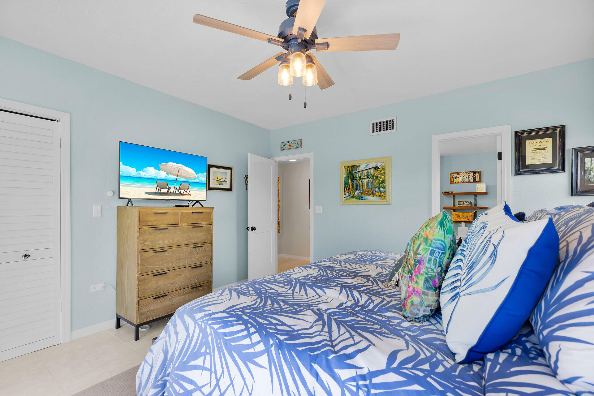 Bedroom with blue-and-white tropical bedding, pale blue walls, and a ceiling fan with lights.