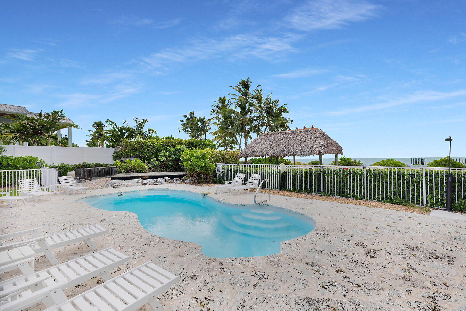Freeform pool with white loungers, a thatched tiki hut, and ocean beyond.