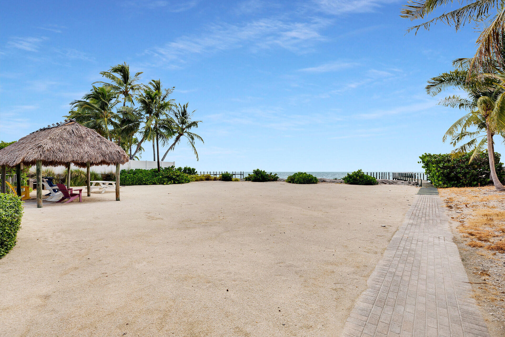 Sandy beachfront with a thatched tiki hut, palm trees, and a paved path to the ocean.