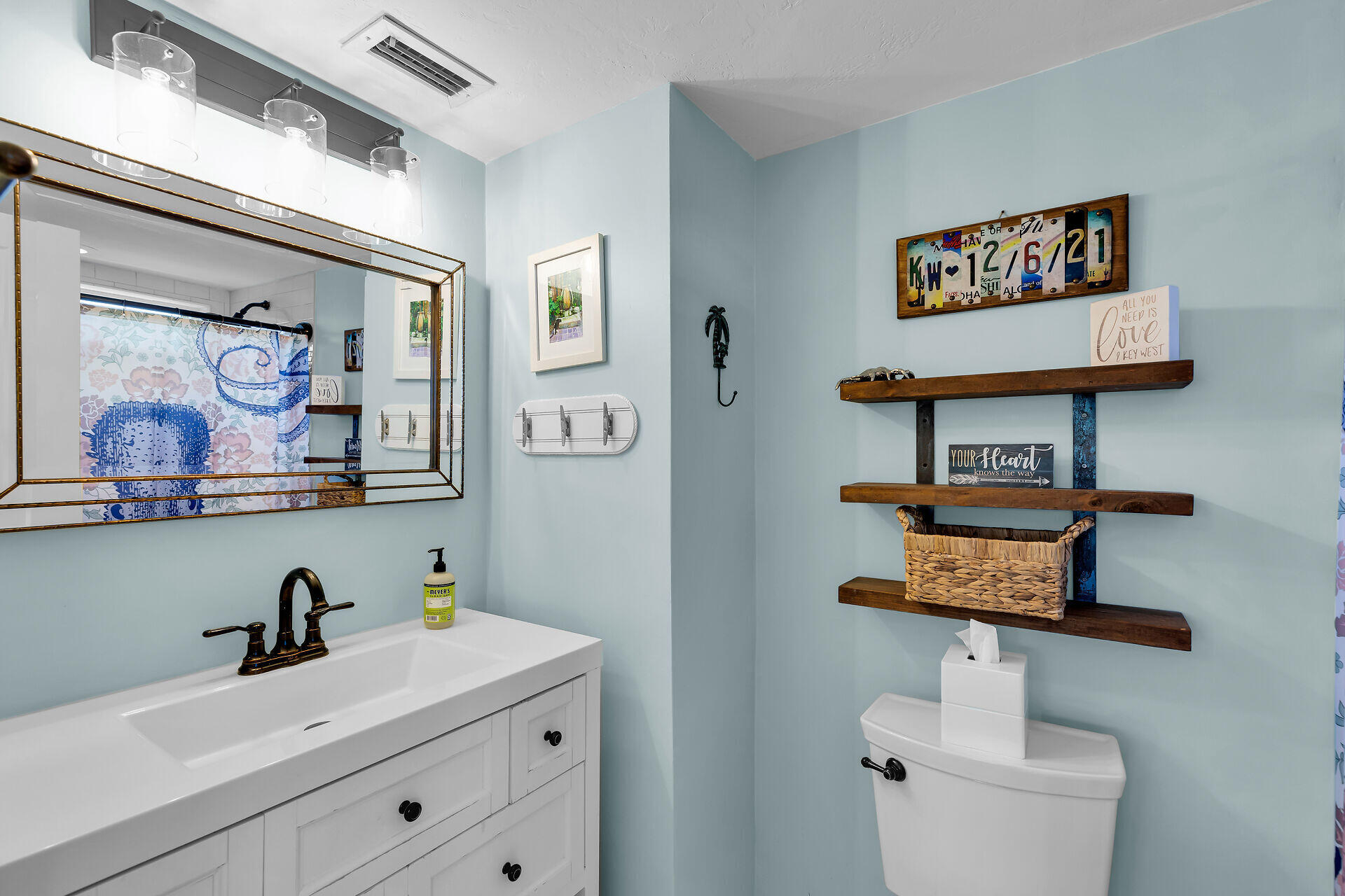 Bathroom with pale blue walls, white vanity, gold-framed mirror, and wooden shelves above the toilet.