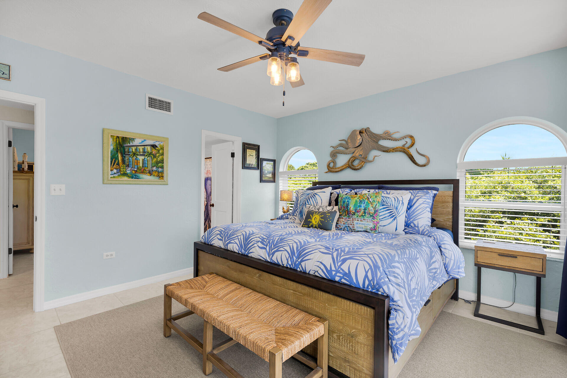 Furnished bedroom with blue palm-print bedding, arched windows, and an octopus wall sculpture.