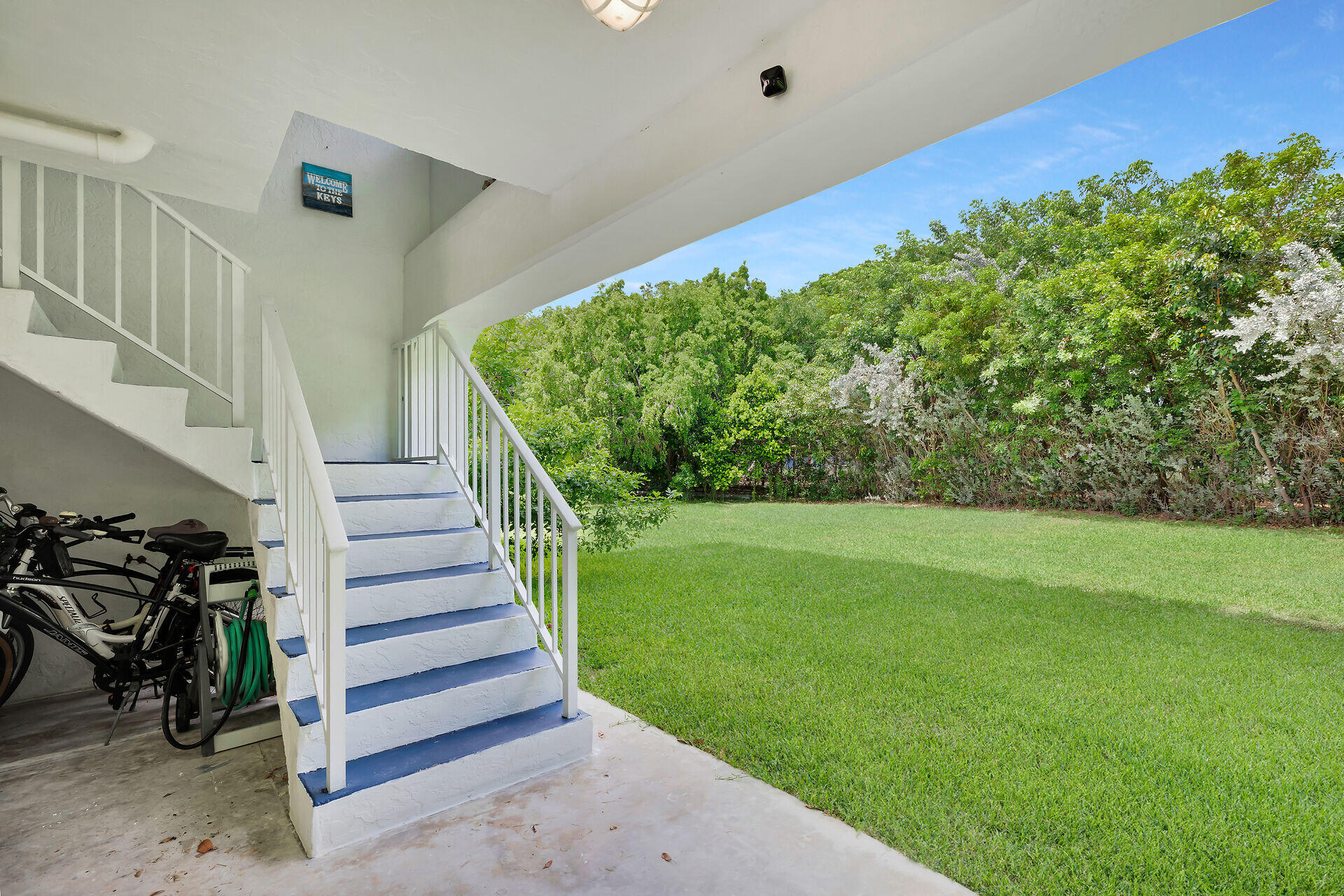 Covered patio with blue-and-white stairs, bicycles, and a lawn bordered by tropical hedges.