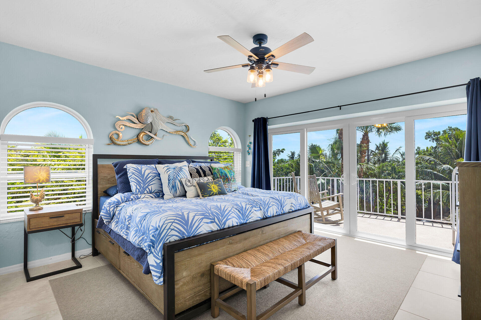 Bedroom with tropical blue bedding, octopus wall art, and sliding doors to a palm-lined balcony.