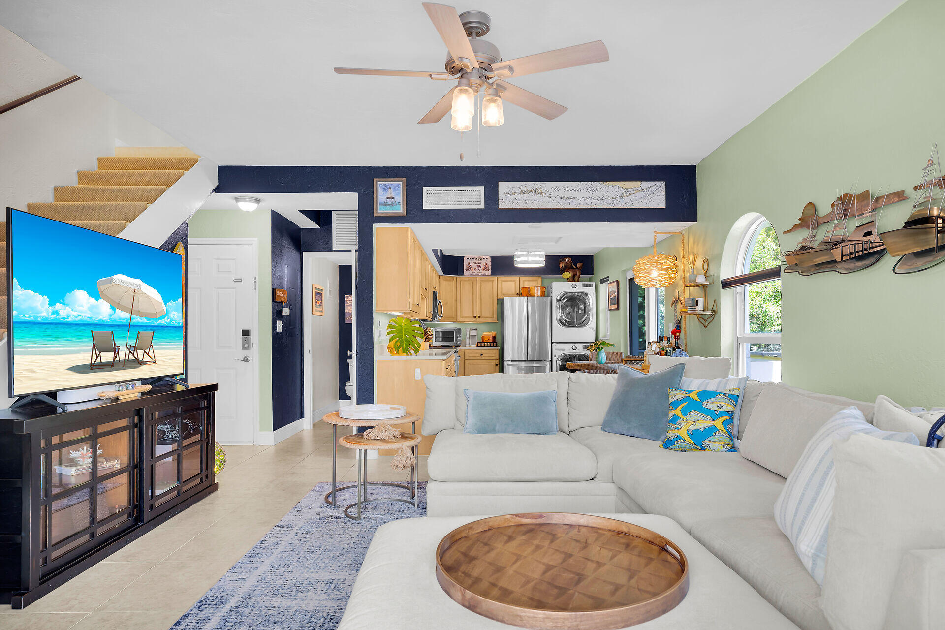 Furnished living room with a white sectional, blue accents, staircase, and open kitchen.