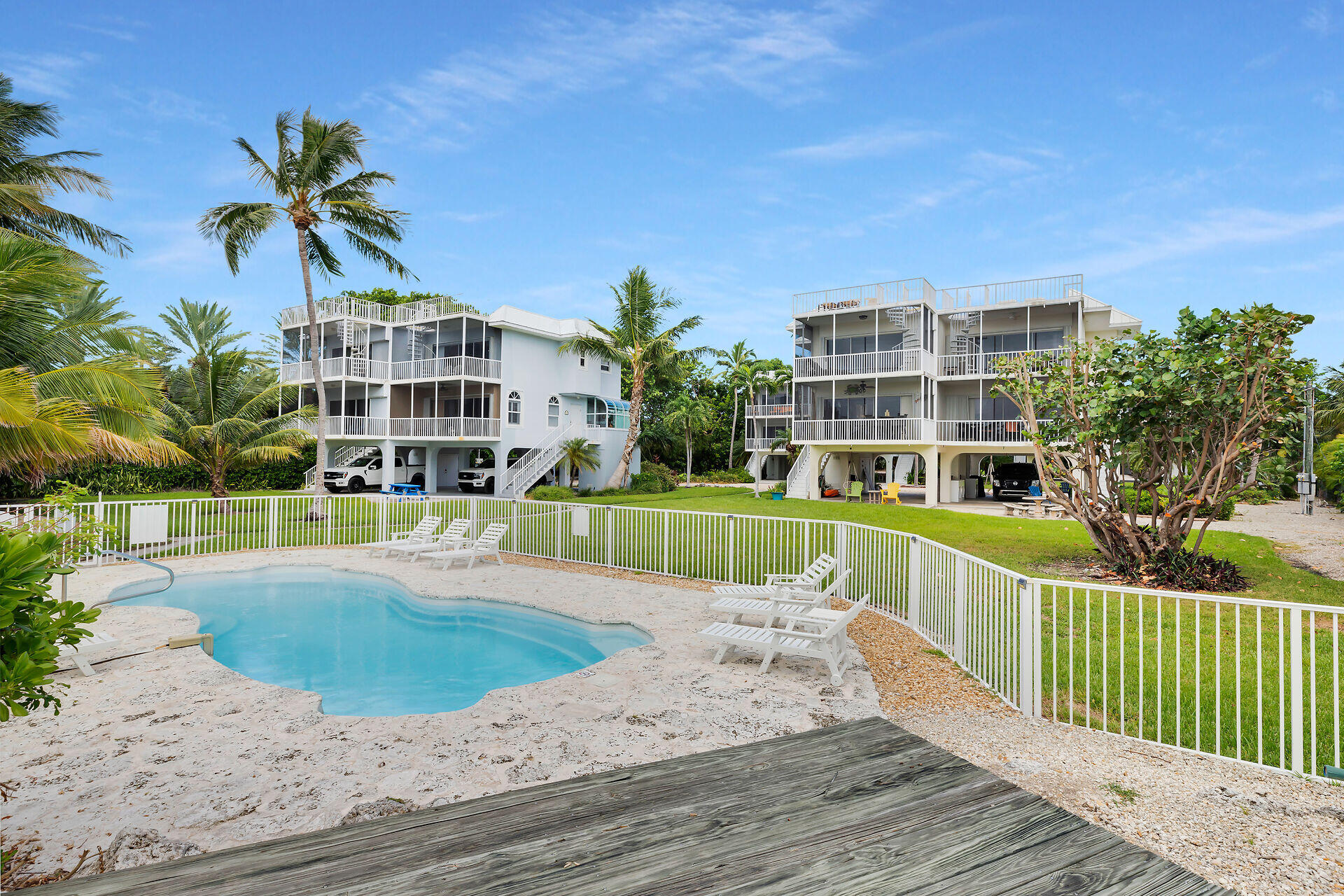 Two elevated white homes with balconies stand beside a fenced pool and palm trees.