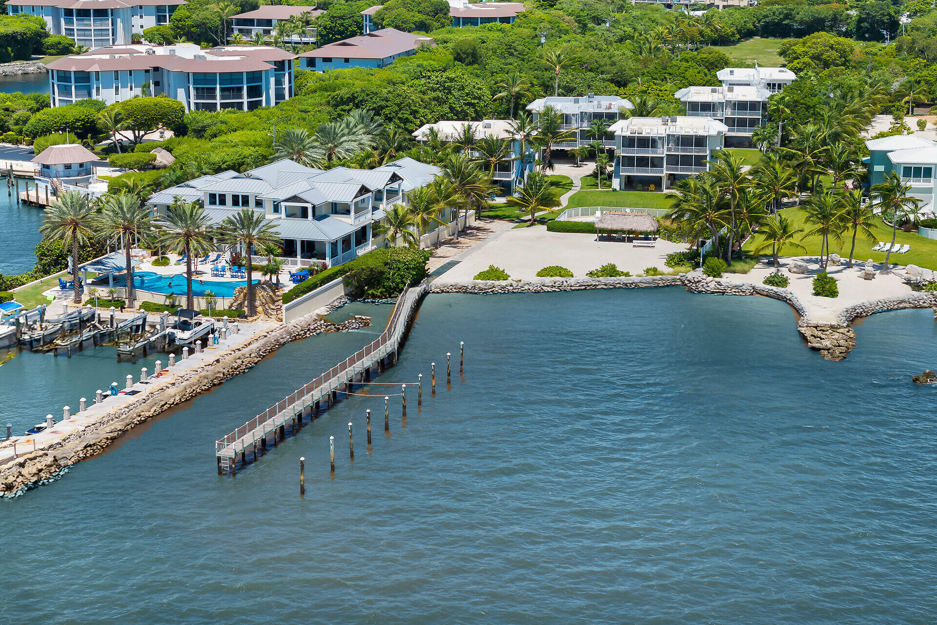 Aerial view of waterfront homes with a pool, docks, palm-lined shore, and surrounding water.