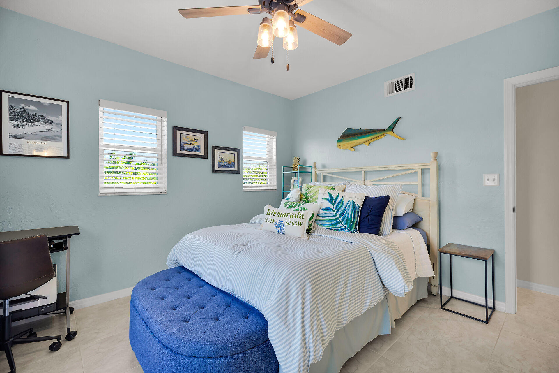 Furnished bedroom with striped bedding, tropical pillows, pale blue walls, and a blue bench.