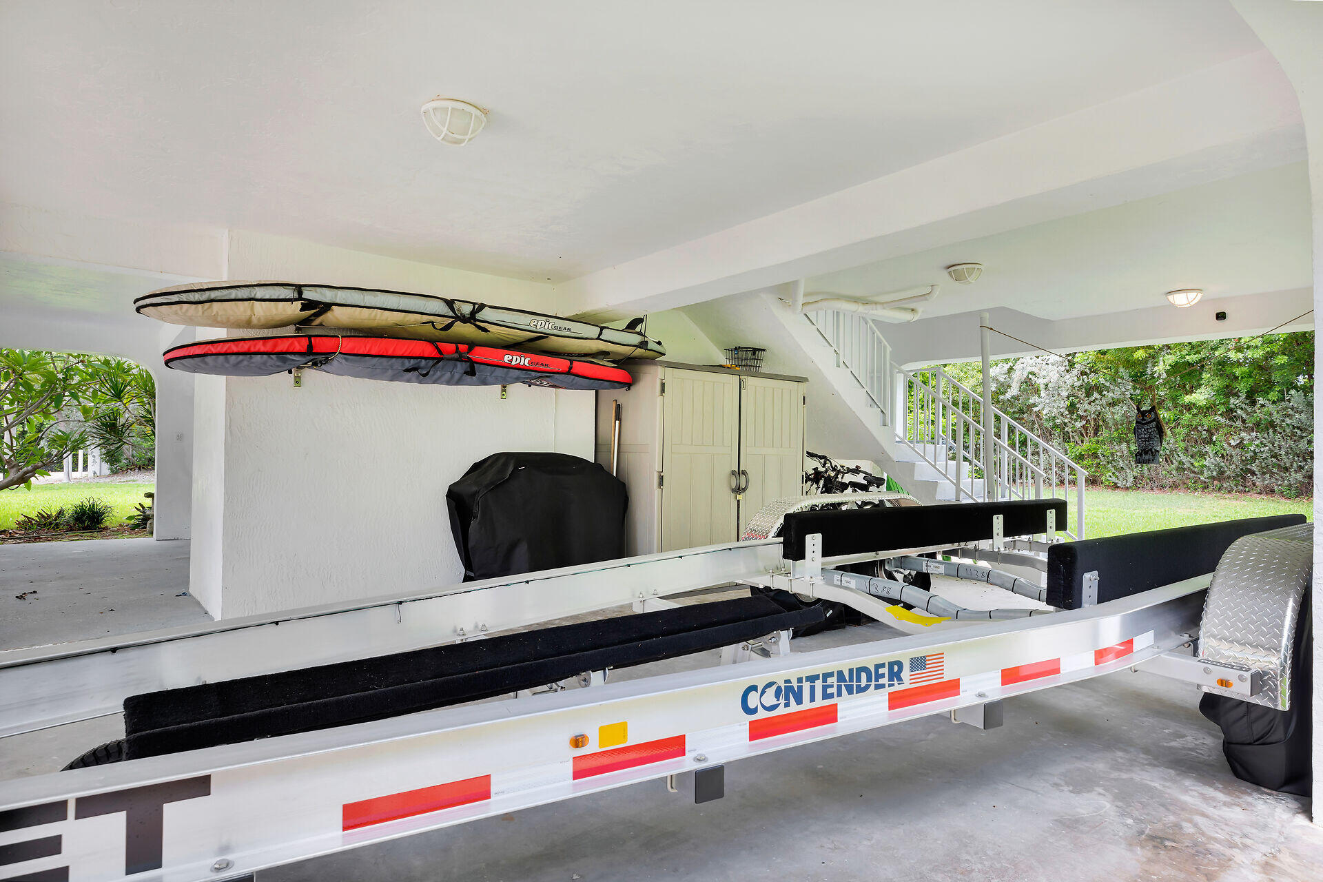 Covered garage with a boat trailer, overhead kayaks, bicycles, and stairs opening onto a leafy yard.