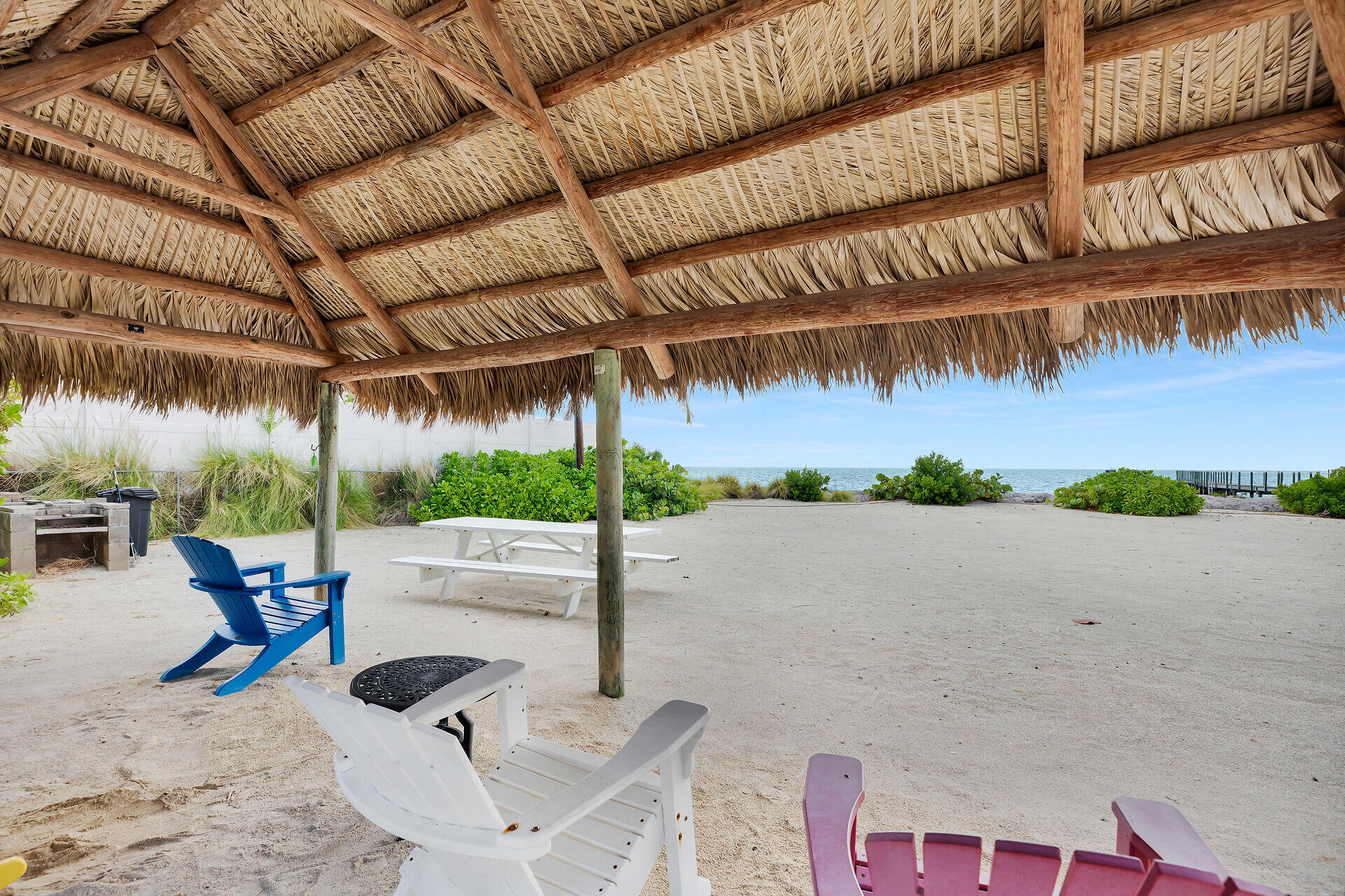 Waterfront sandy patio beneath a thatched tiki pavilion, with colorful chairs, ocean, and dock.