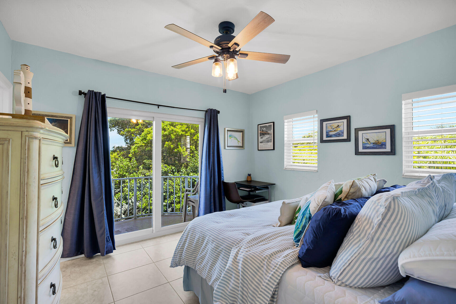 Furnished bedroom with blue-and-white bedding, sliding balcony doors, and leafy greenery outside.