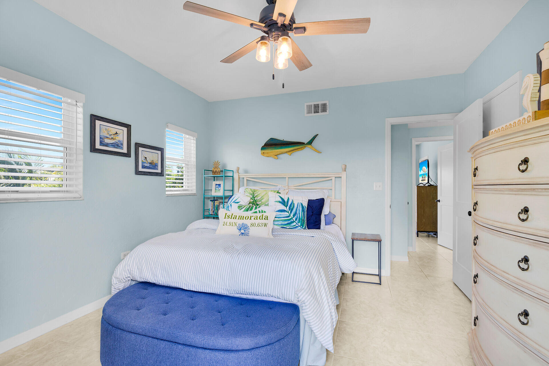 Bedroom with pale blue walls, striped bedding, and a blue bench beneath coastal fish decor.