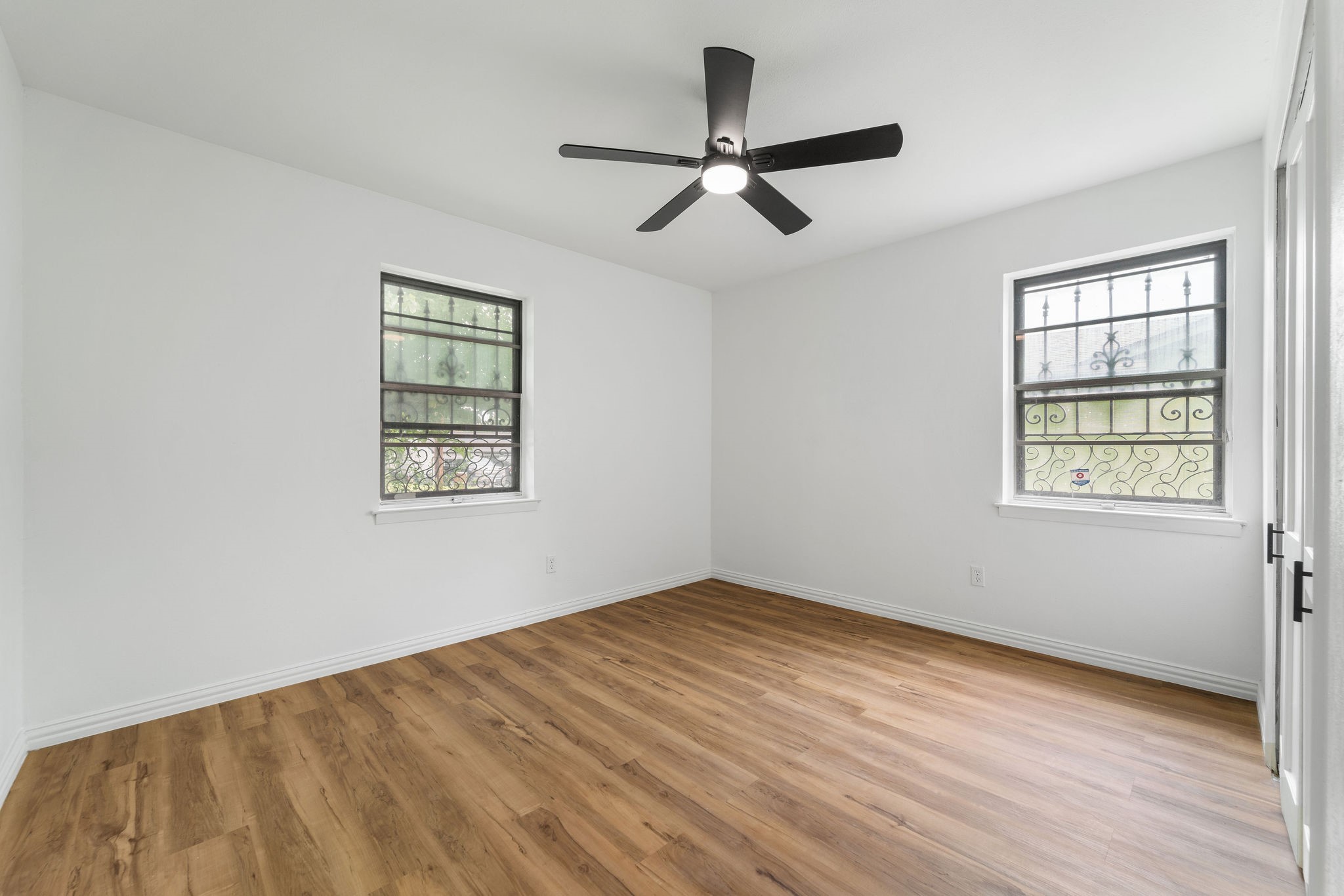 Unfurnished room with wood-look floors, a black ceiling fan, and two windows with decorative grilles.