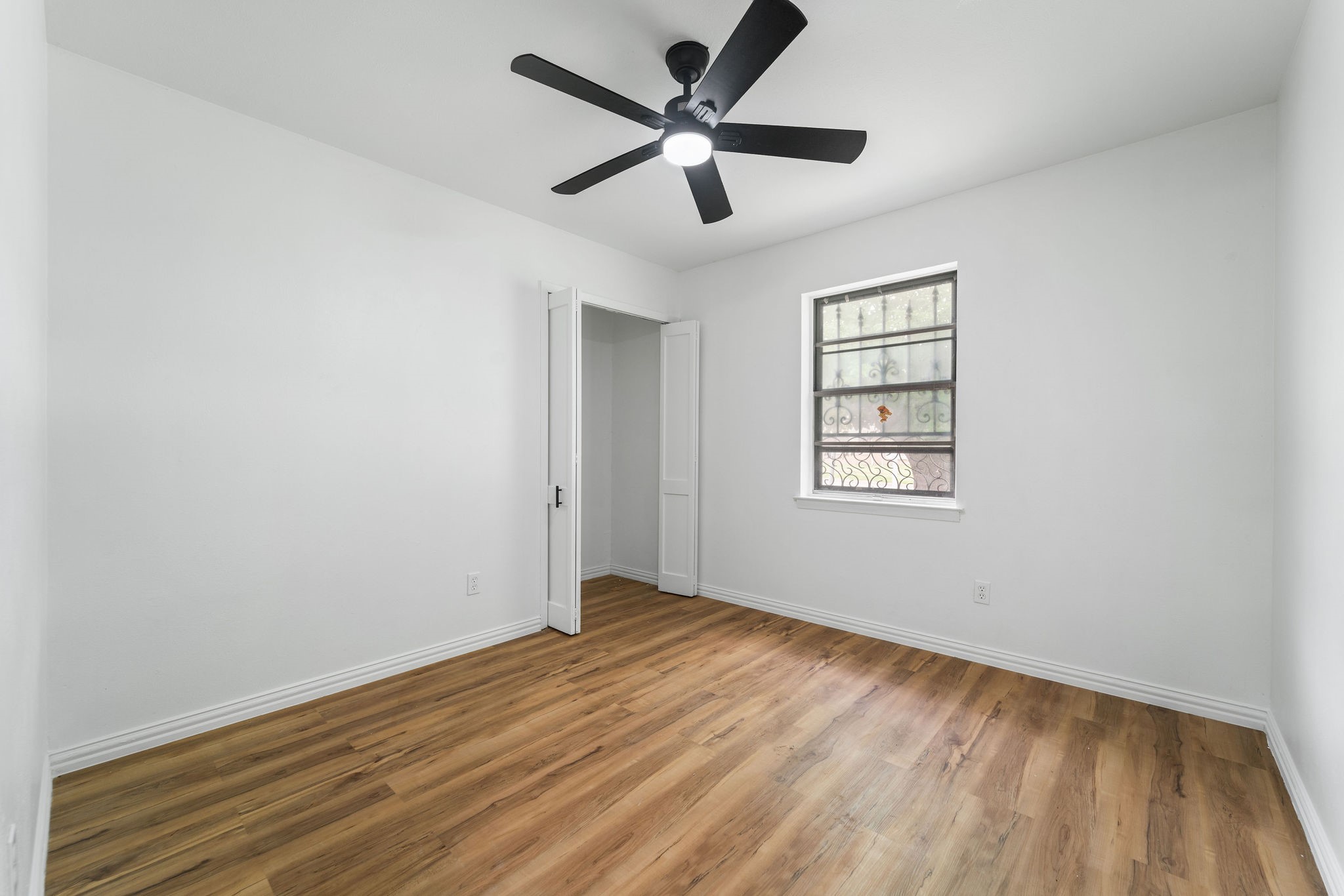 Empty bedroom with wood-look floors, a black ceiling fan, and a barred window.