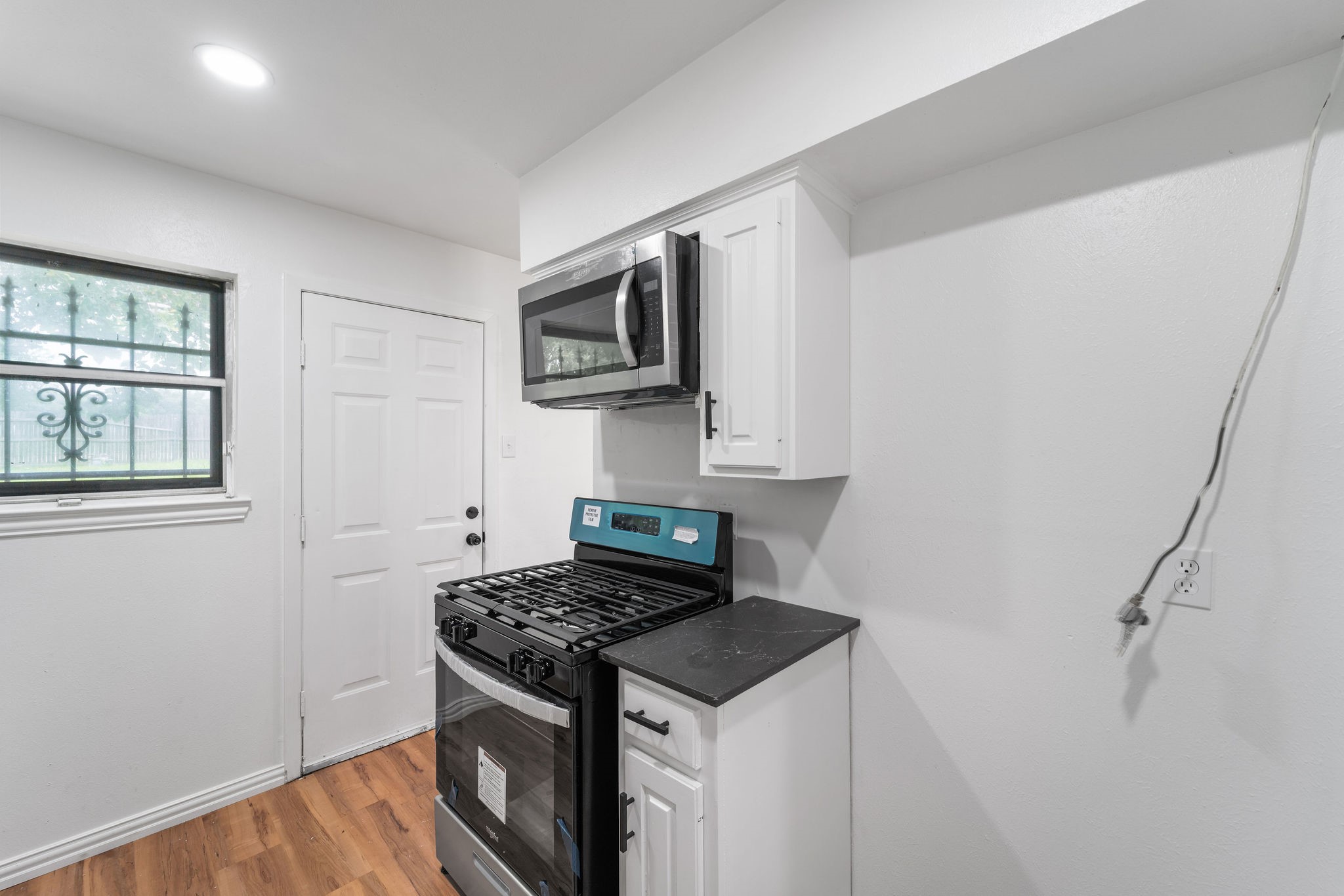 Compact kitchen with a black gas range, over-the-range microwave, white cabinets, and a barred window.