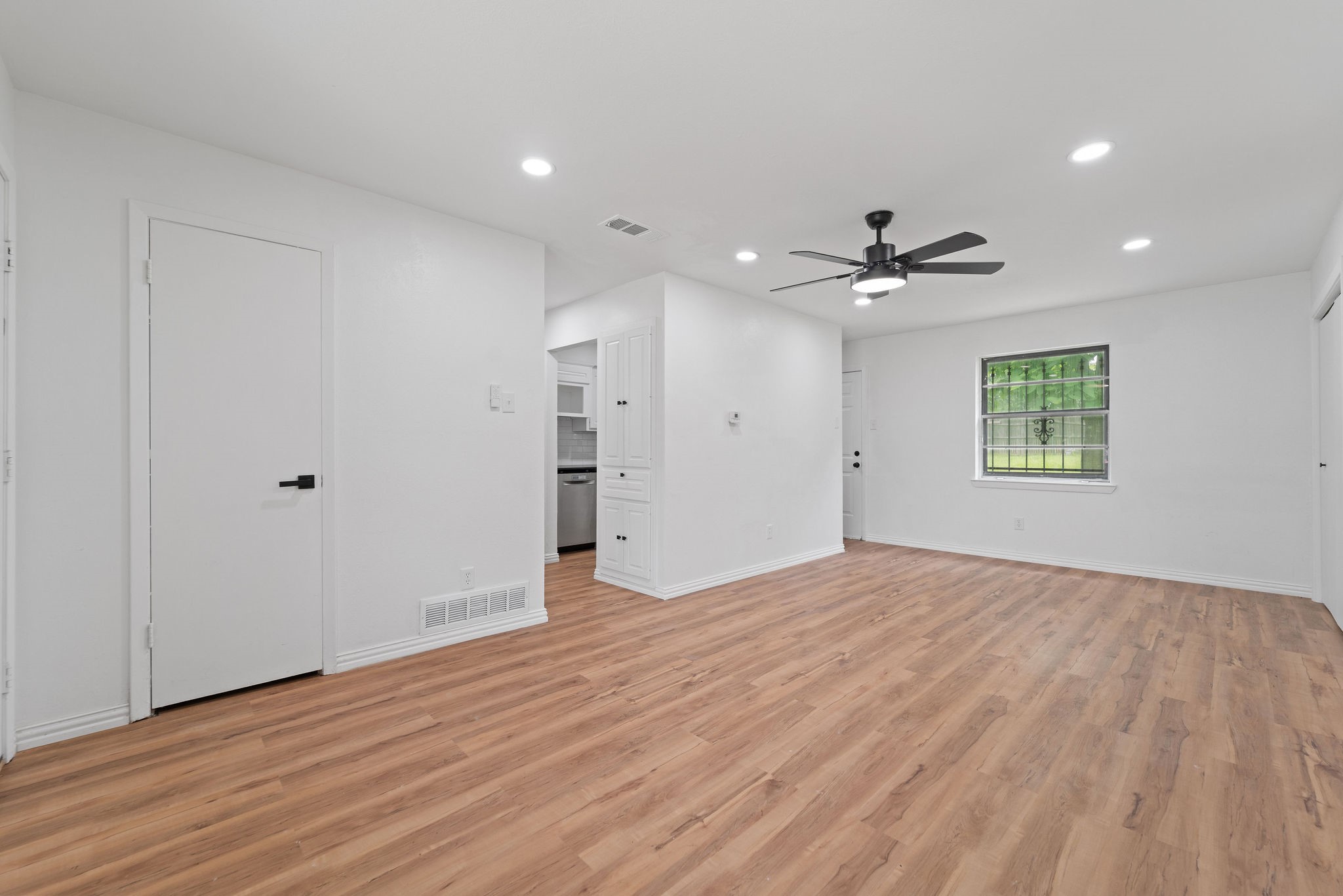 Unfurnished living room with warm wood flooring, a dark ceiling fan, and a window overlooking greenery.
