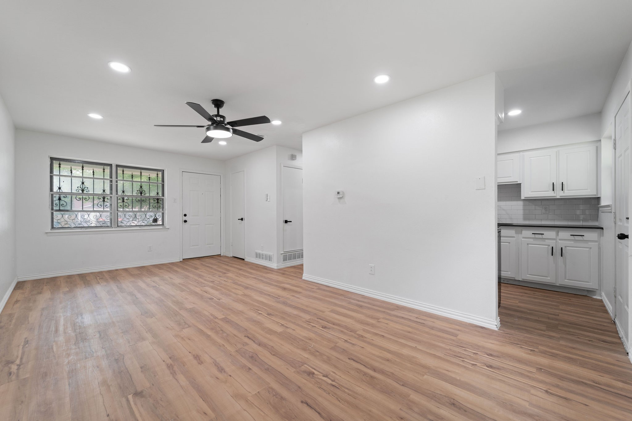 Vacant living room with wood-look floors, a black ceiling fan, and an adjoining white kitchen.