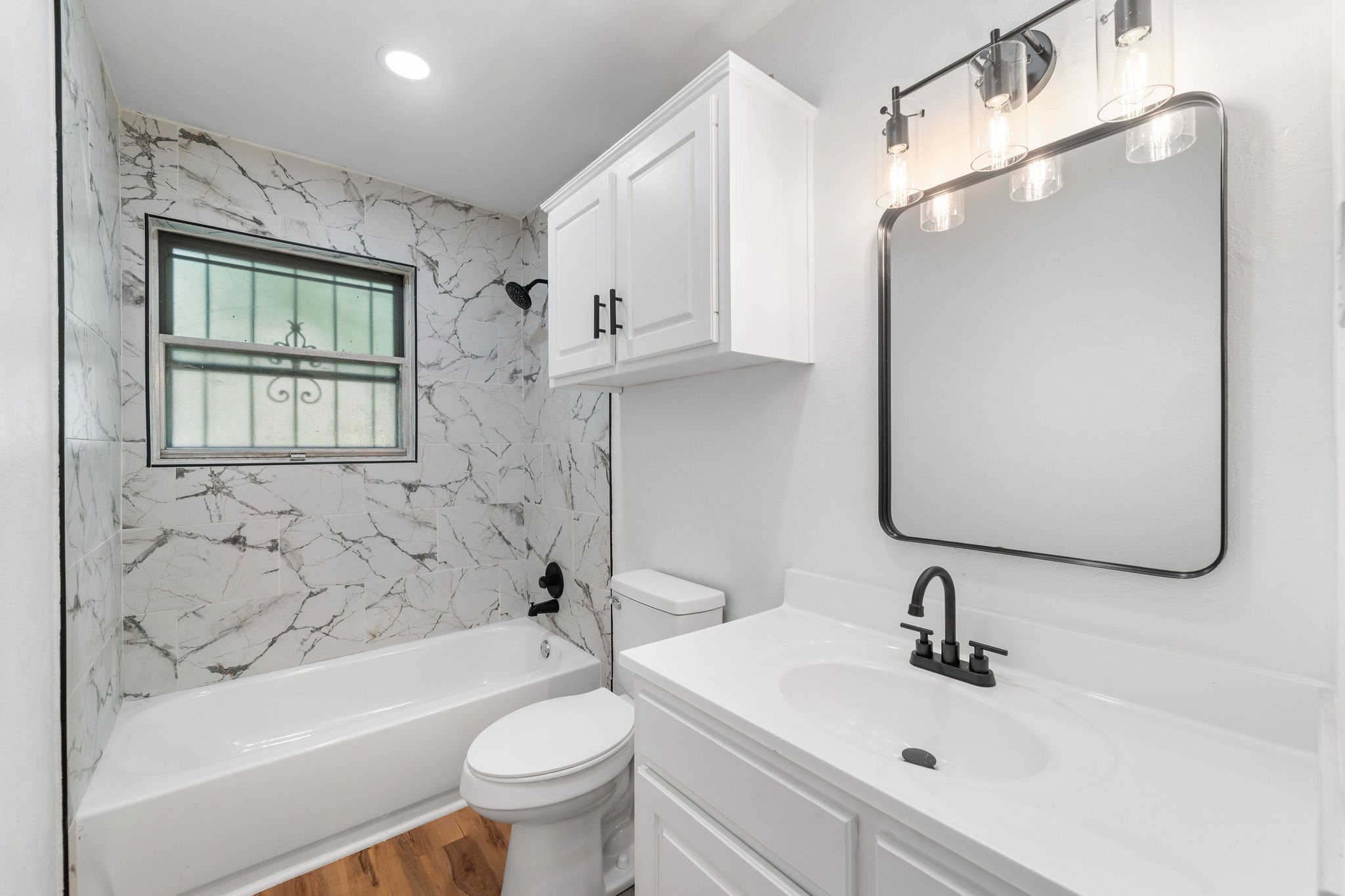 Bathroom with marble-look tub surround, white vanity, and black fixtures.