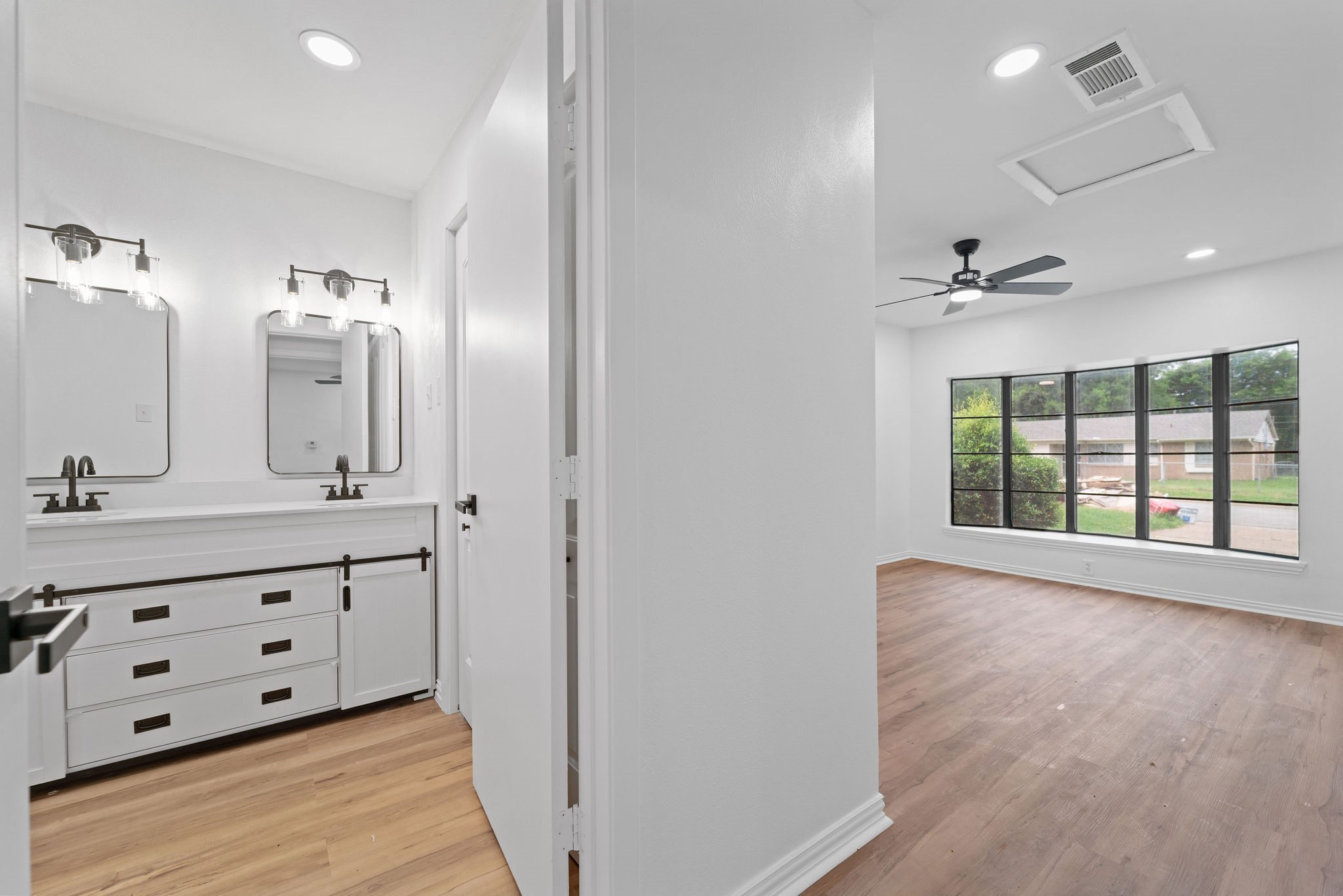 Bathroom with a white double vanity and dark fixtures, opening onto a bright empty room with large windows.