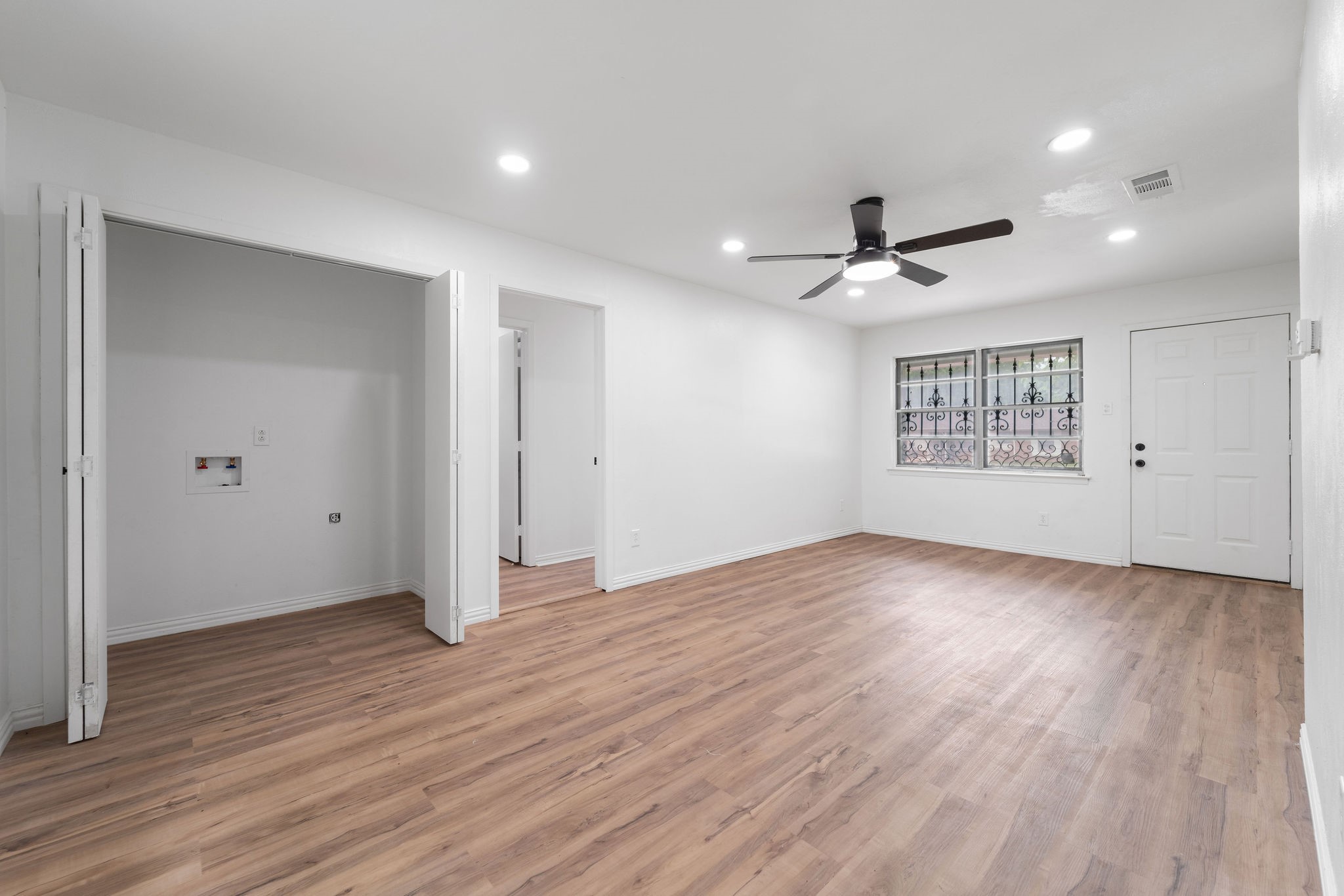 Vacant room with wood-look floors, a ceiling fan, and a barred window beside the front door.