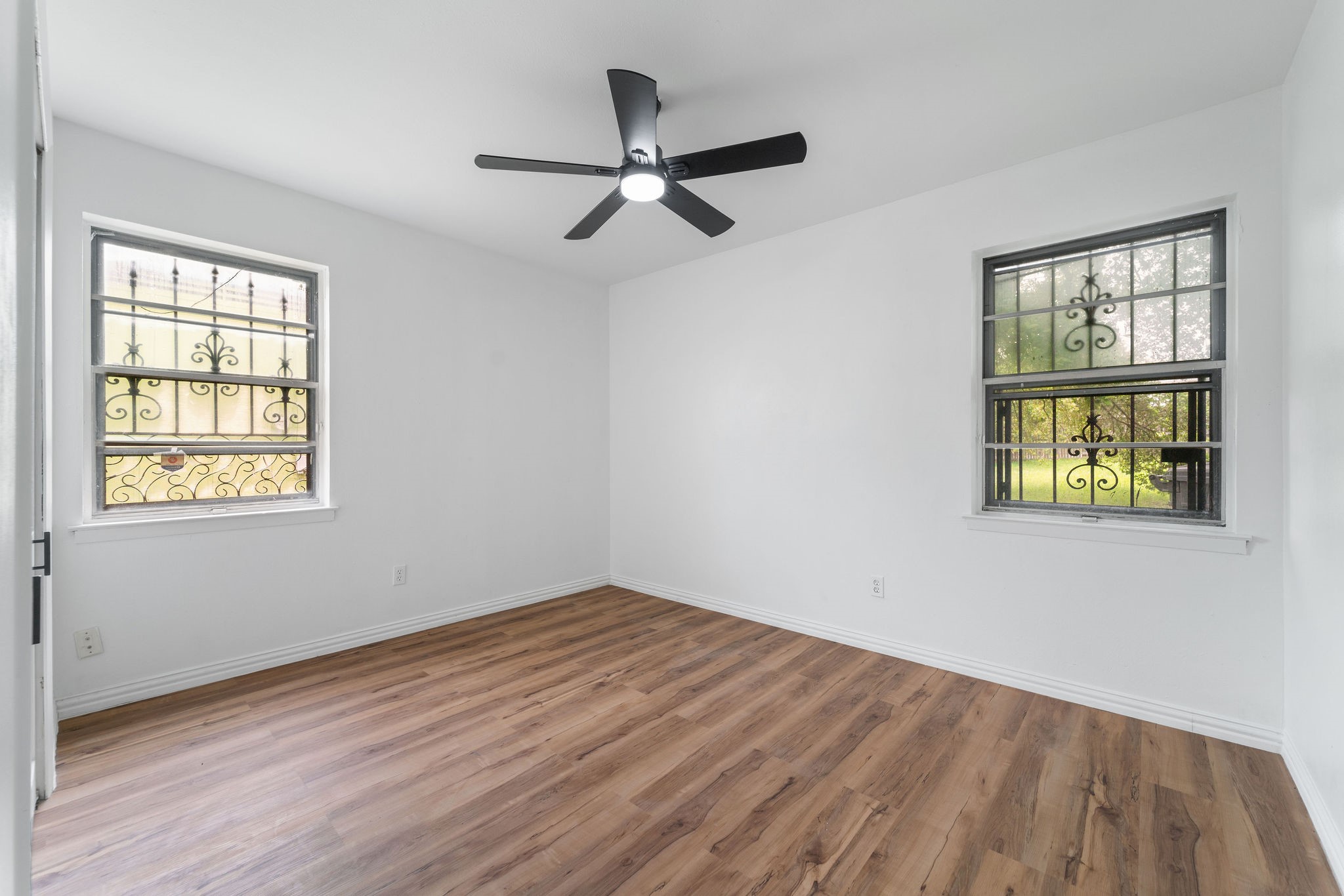 Empty room with wood-look flooring, a ceiling fan, and two windows with decorative metal bars.