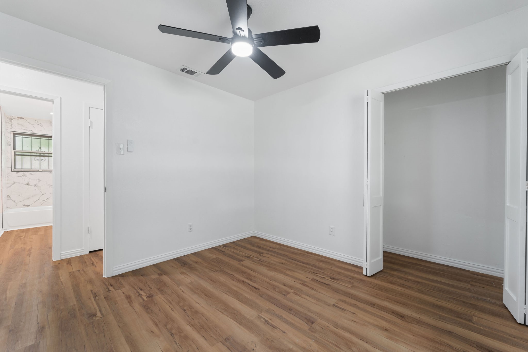Empty bedroom with wood-look floors, a black ceiling fan, and open closet doors.