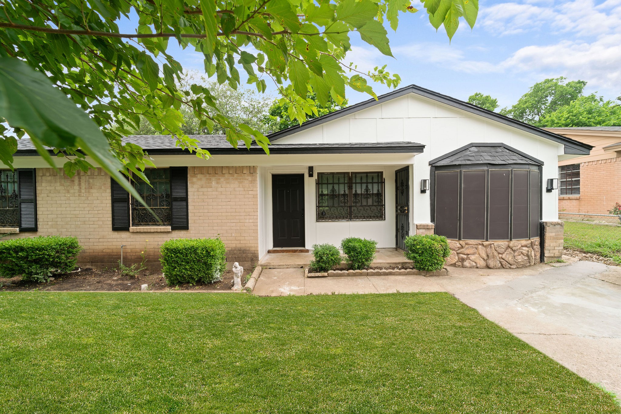 Single-story house with tan brick, white siding, a leafy tree overhead, and a green front lawn.