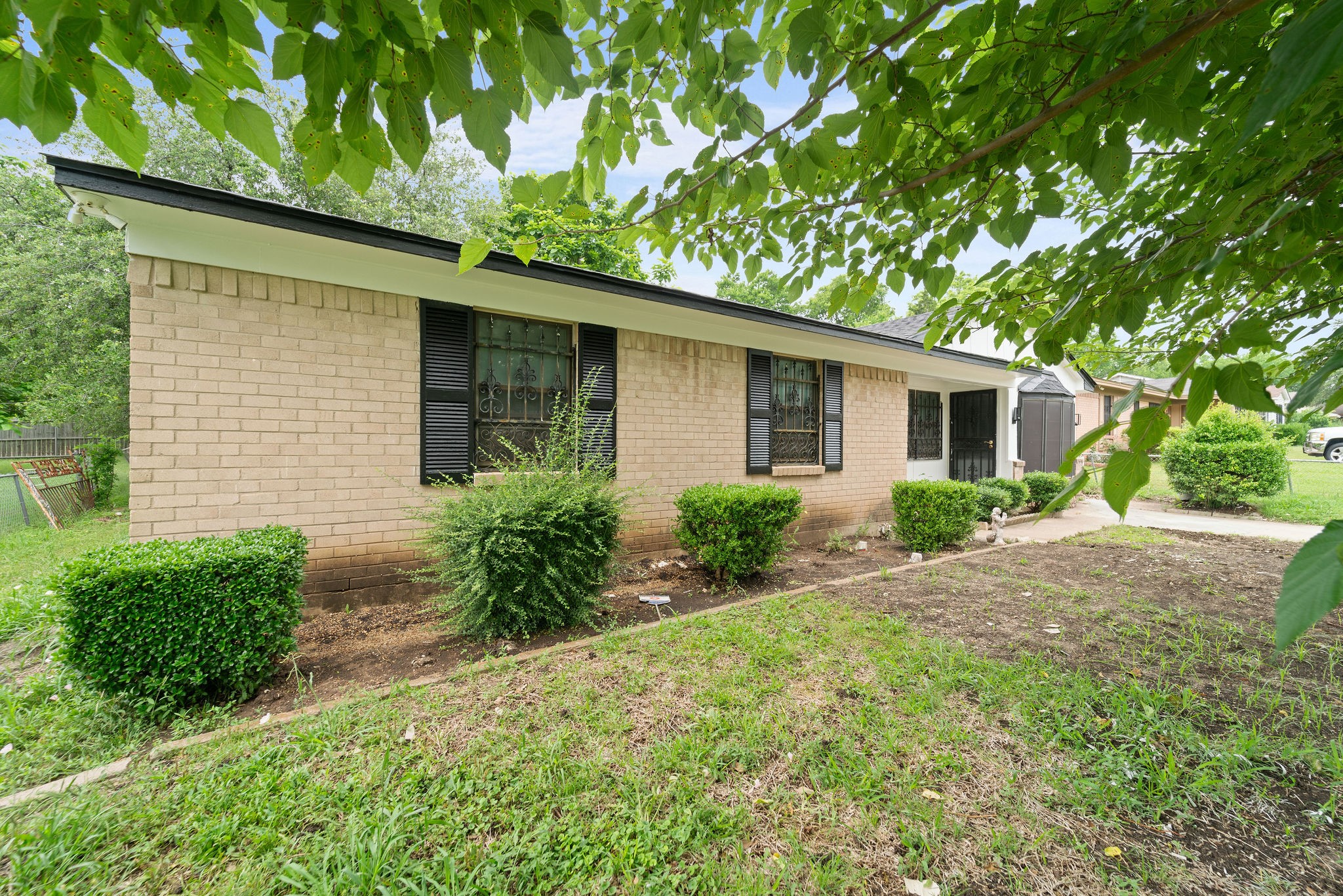 Single-story tan brick home with black shutters and leafy branches over the front yard.