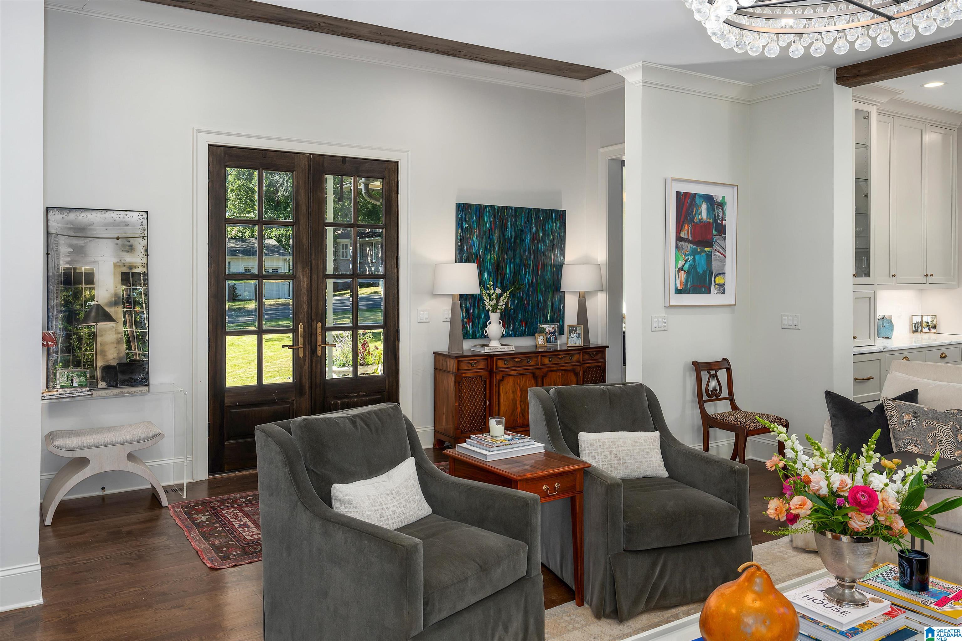 Living room with dark wood French doors, gray armchairs, exposed beams, and a crystal chandelier.