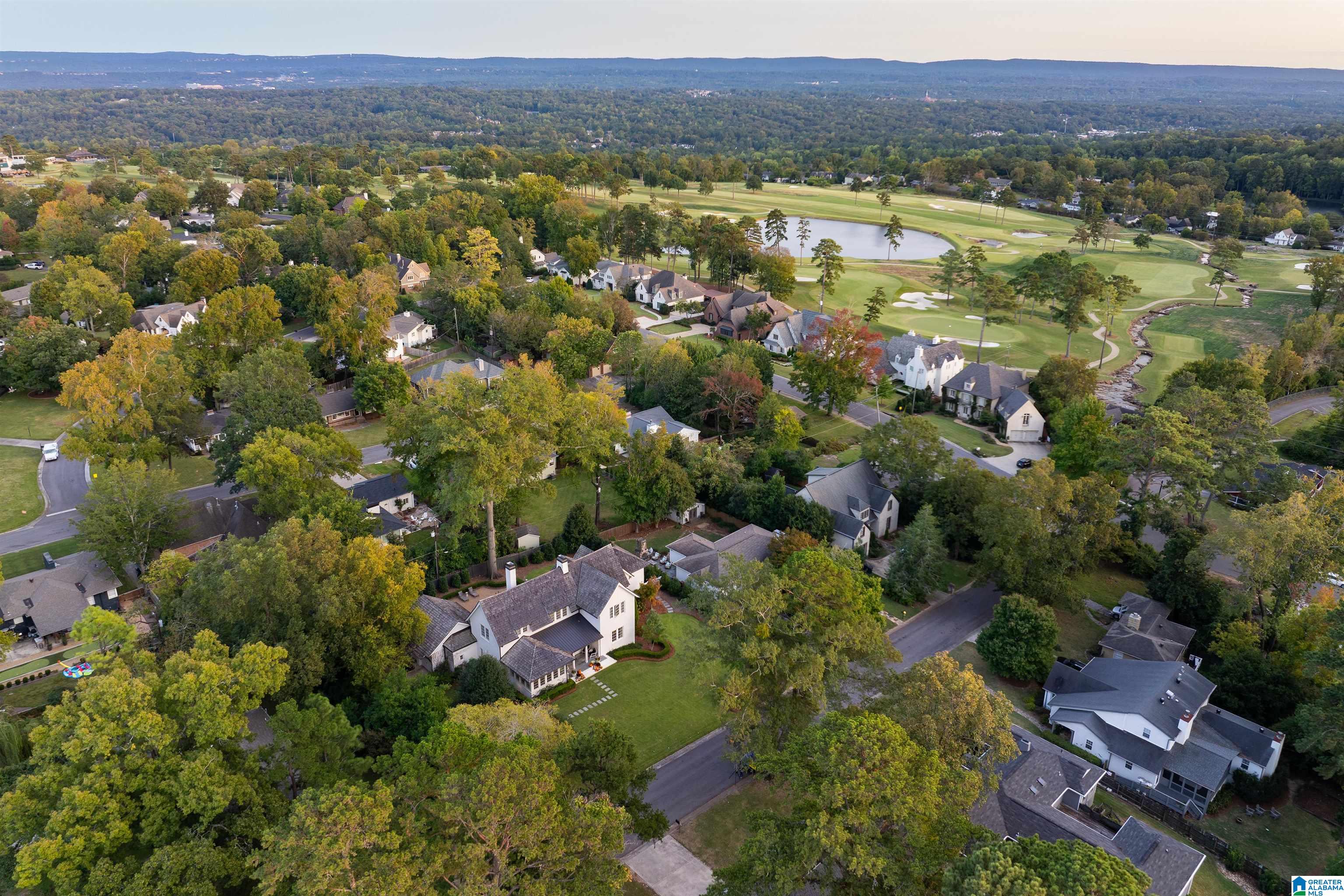 Aerial neighborhood view with tree-lined homes and a golf course, pond, and sand bunkers beyond.