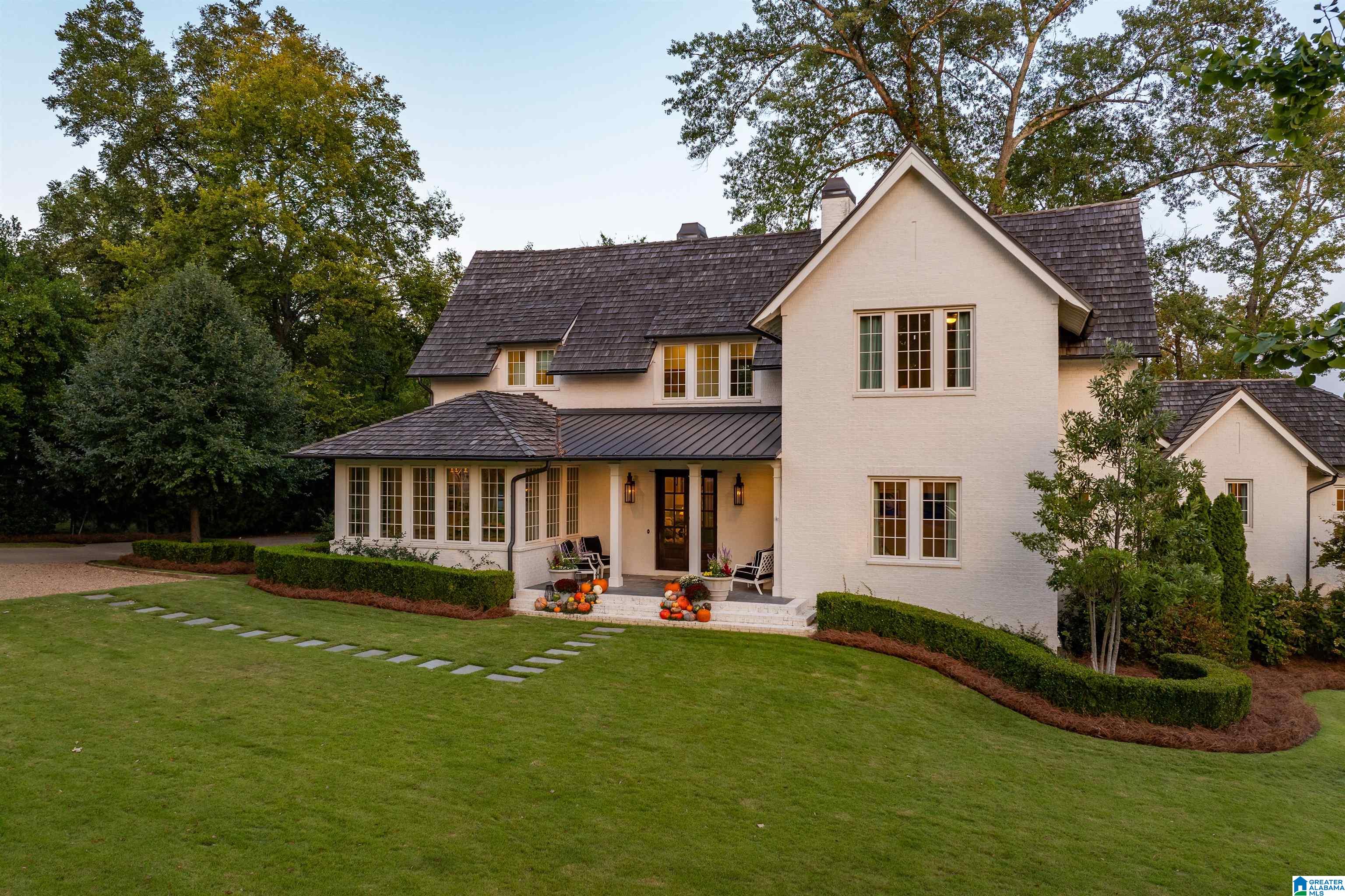 White brick home with dark gabled roofs, a covered porch, broad lawn, and pumpkins on the front steps.