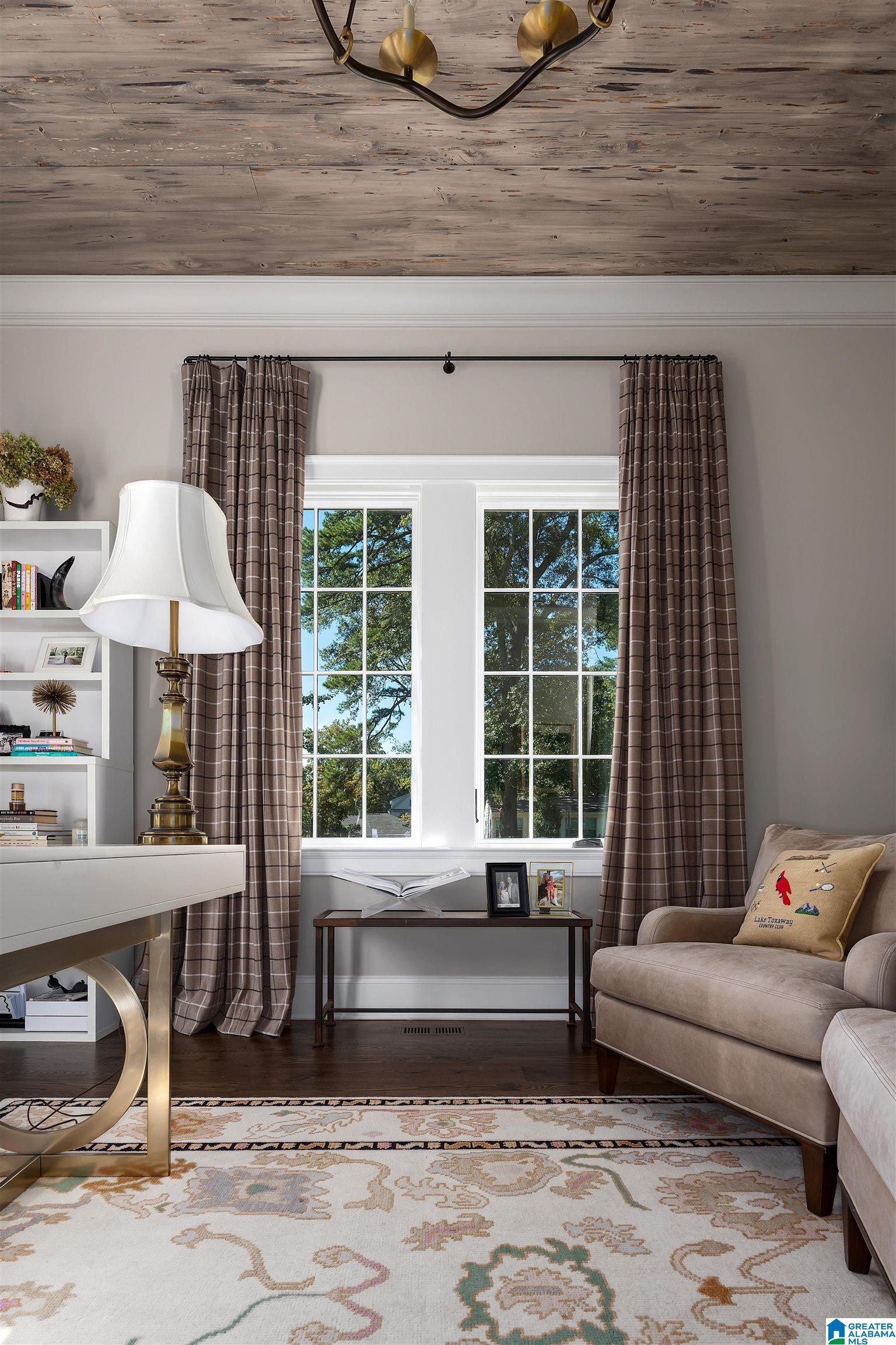 Furnished sitting area with a tall curtained window, beige armchair, white desk, and floral rug.