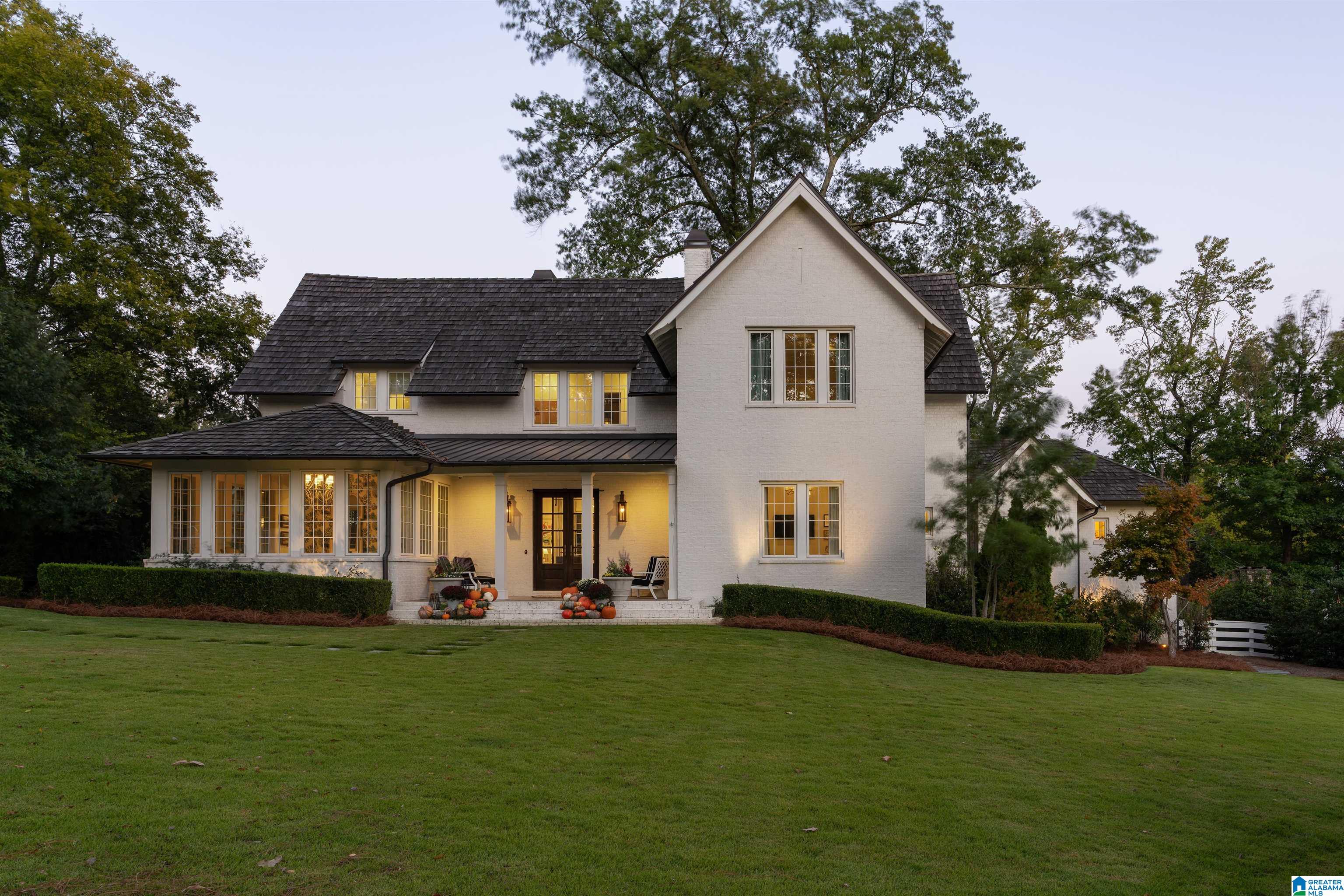 White two-story house with a dark shingle roof, broad lawn, and pumpkin-decorated entrance at dusk.