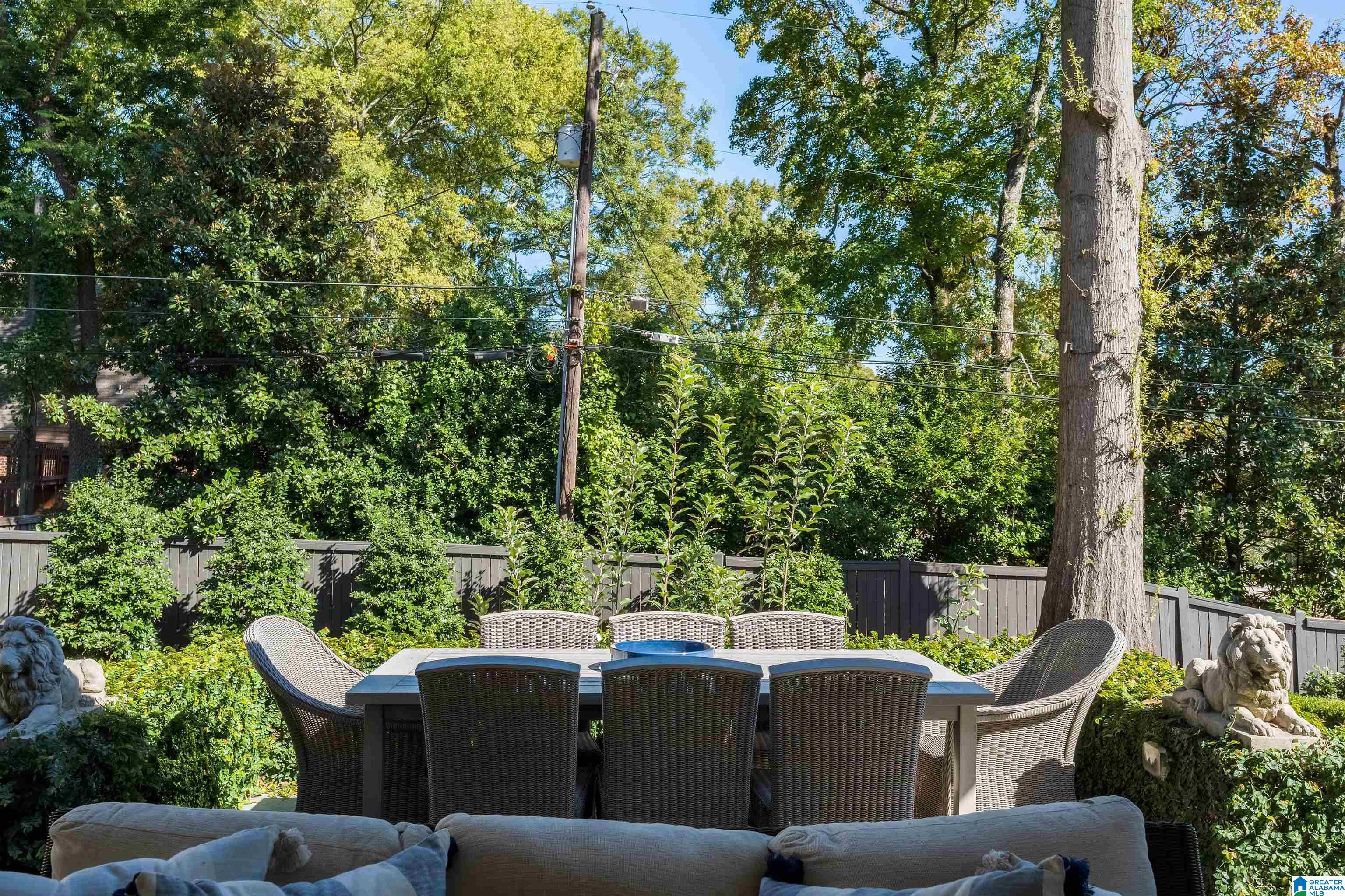 Furnished patio with a woven dining set, cushioned sofa, lush landscaping, and mature trees.