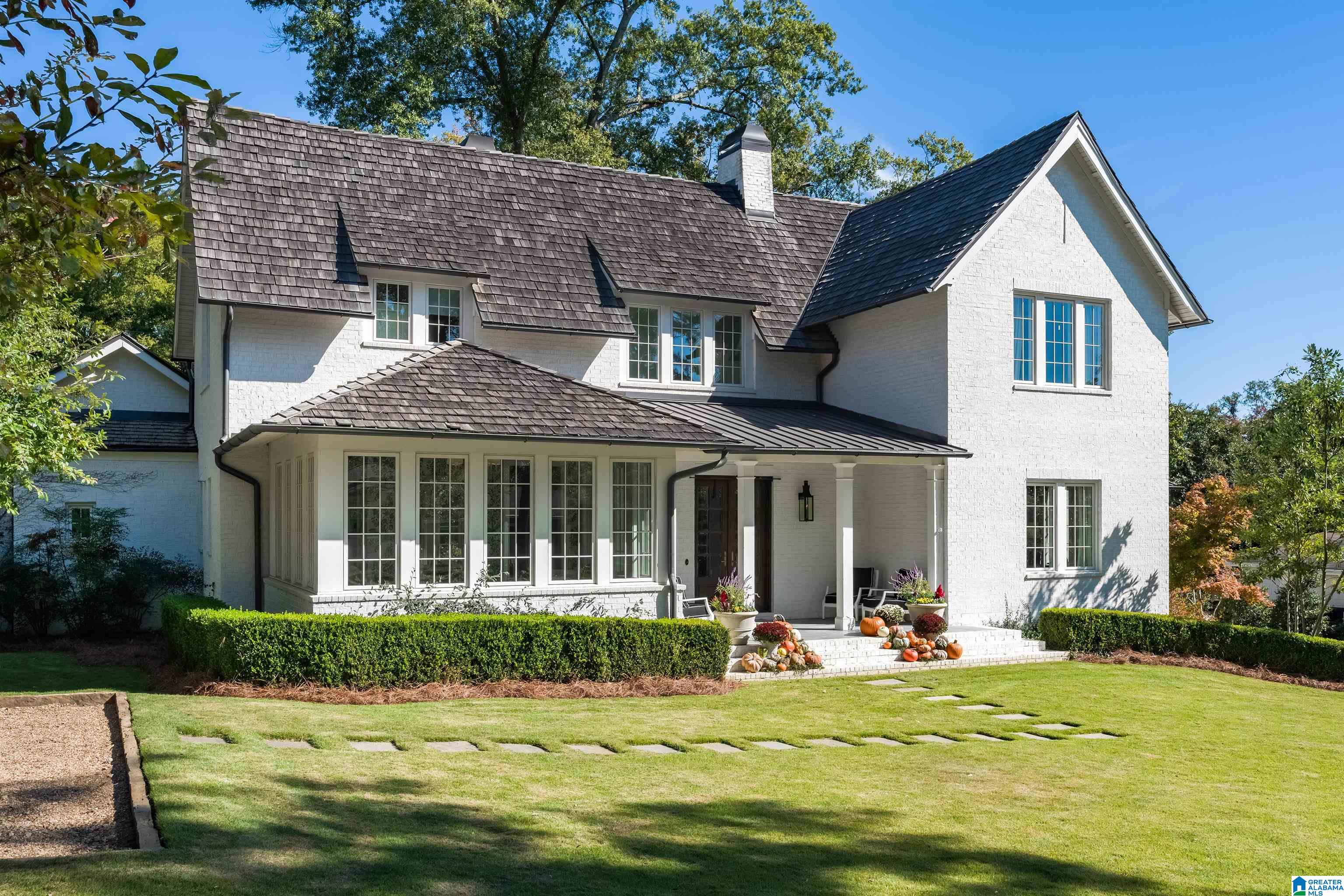 White brick two-story home with dark gabled roofs, a covered porch, and pumpkins on the front steps.