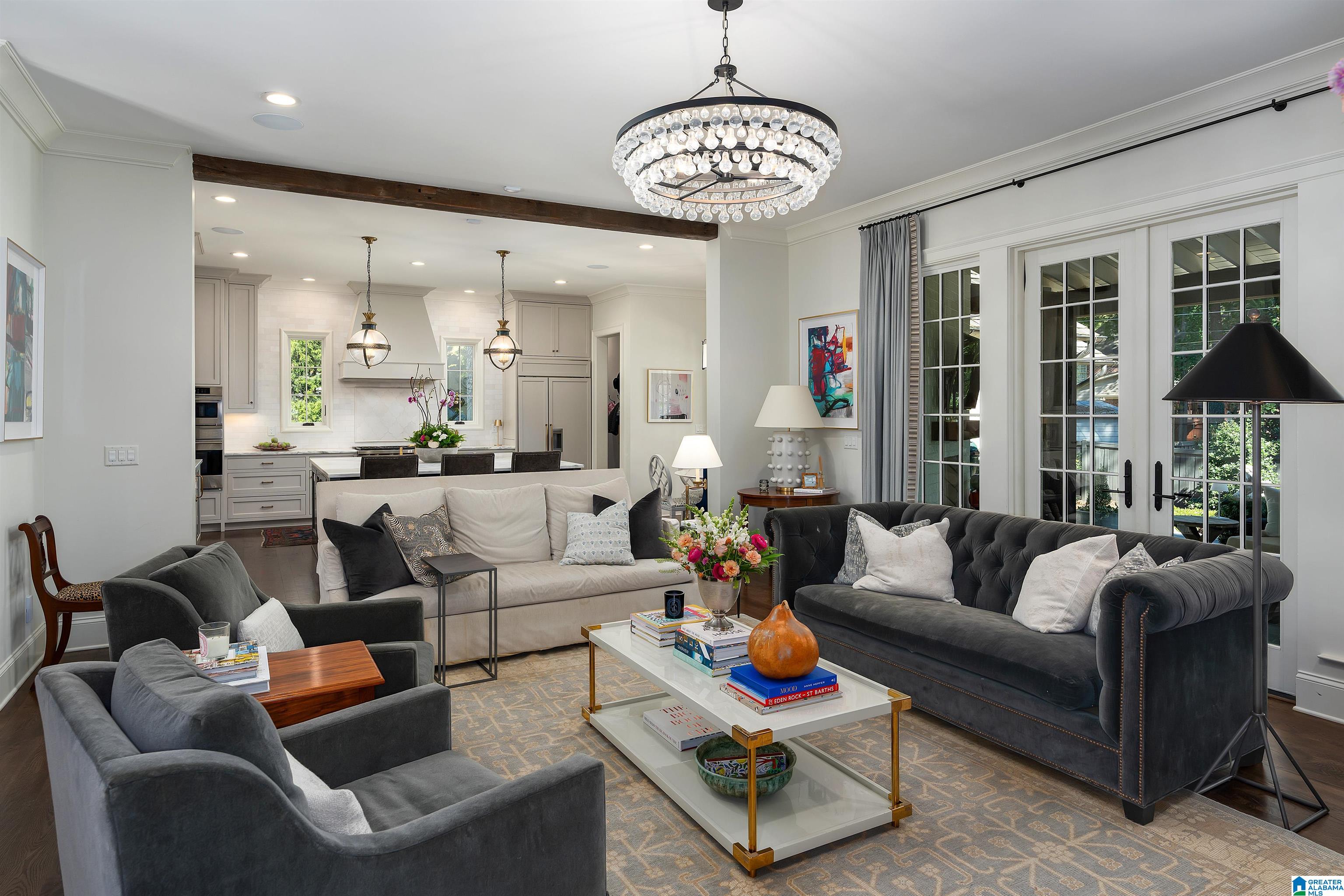 Open-plan living room with a crystal chandelier, gray sofas, French doors, and the kitchen beyond.