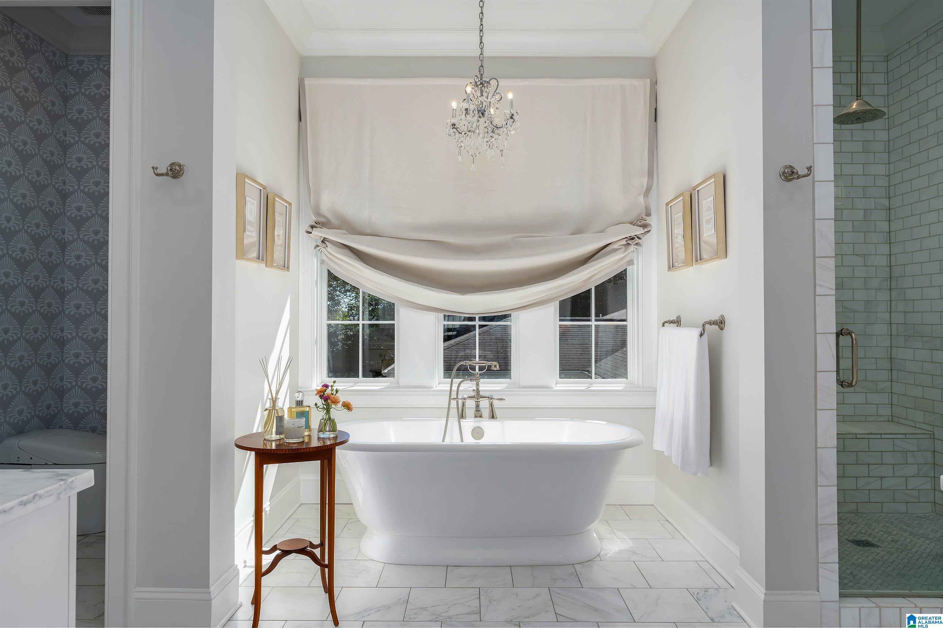 Freestanding soaking tub beneath a curtained bay window, with a chandelier and tiled shower.
