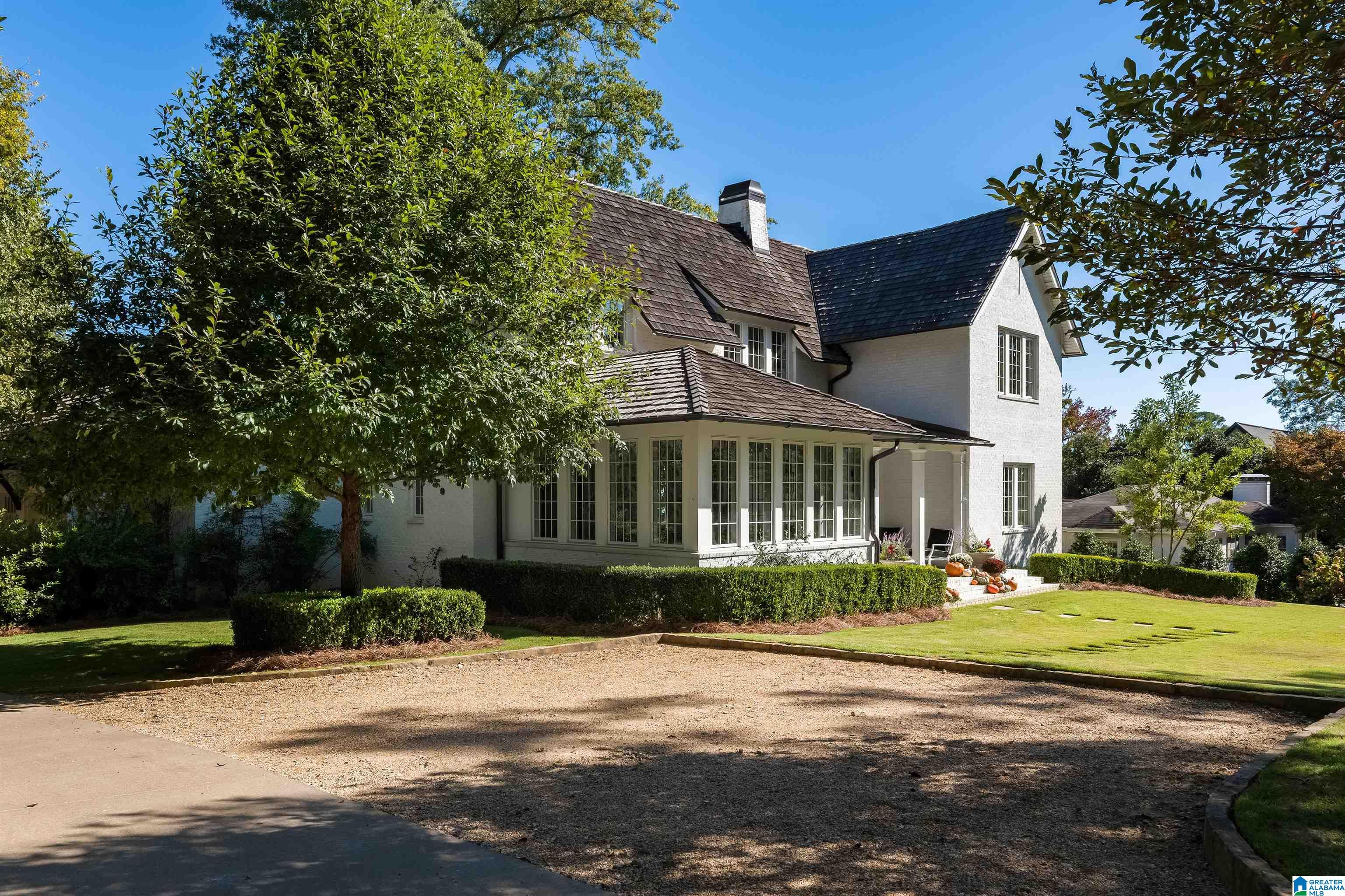 White two-story house with a windowed sunroom, landscaped lawn, gravel drive, and mature trees.