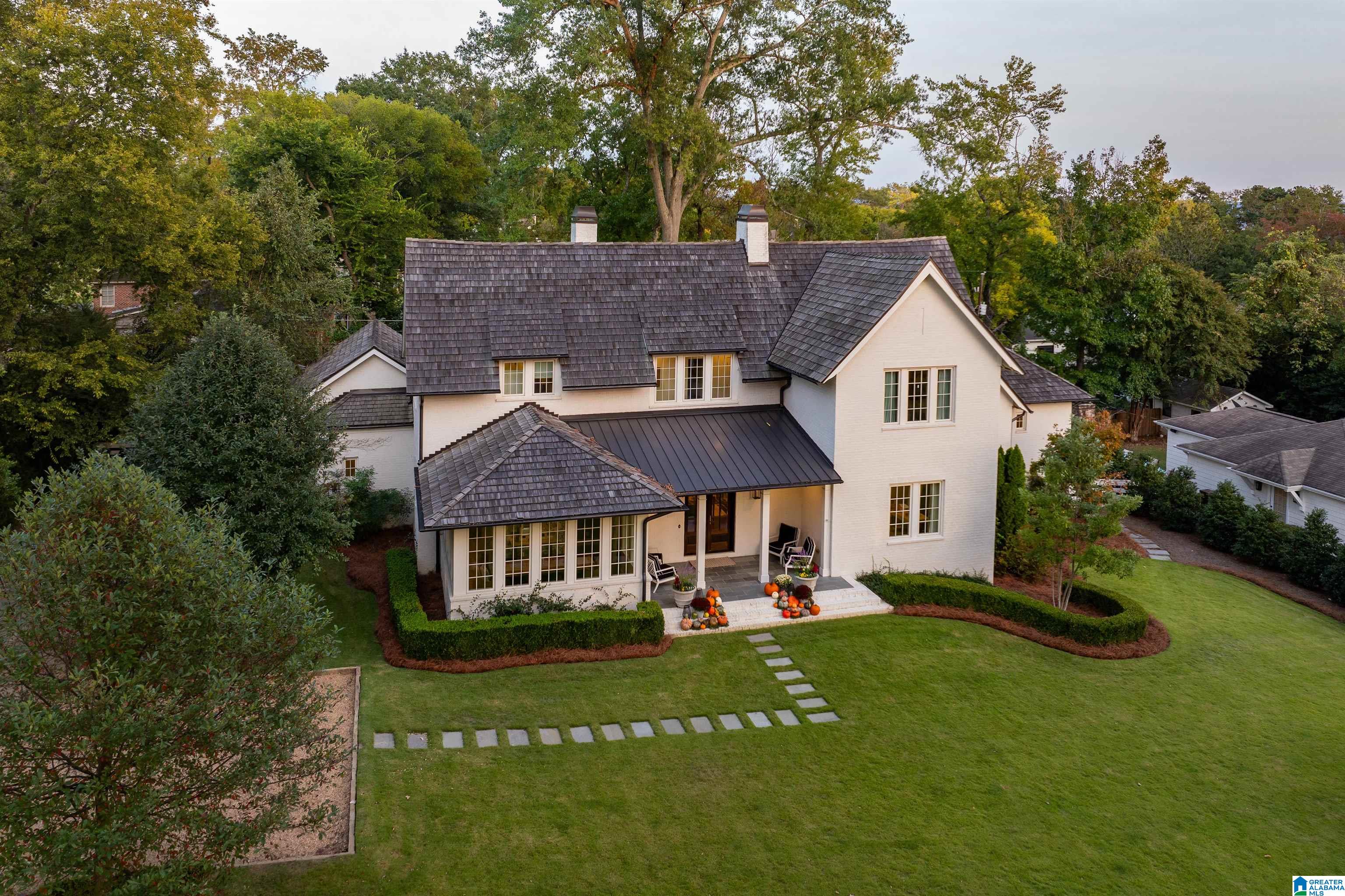 Aerial view of a white two-story house with dark shingle roofs, a green lawn, and pumpkins by the entry.