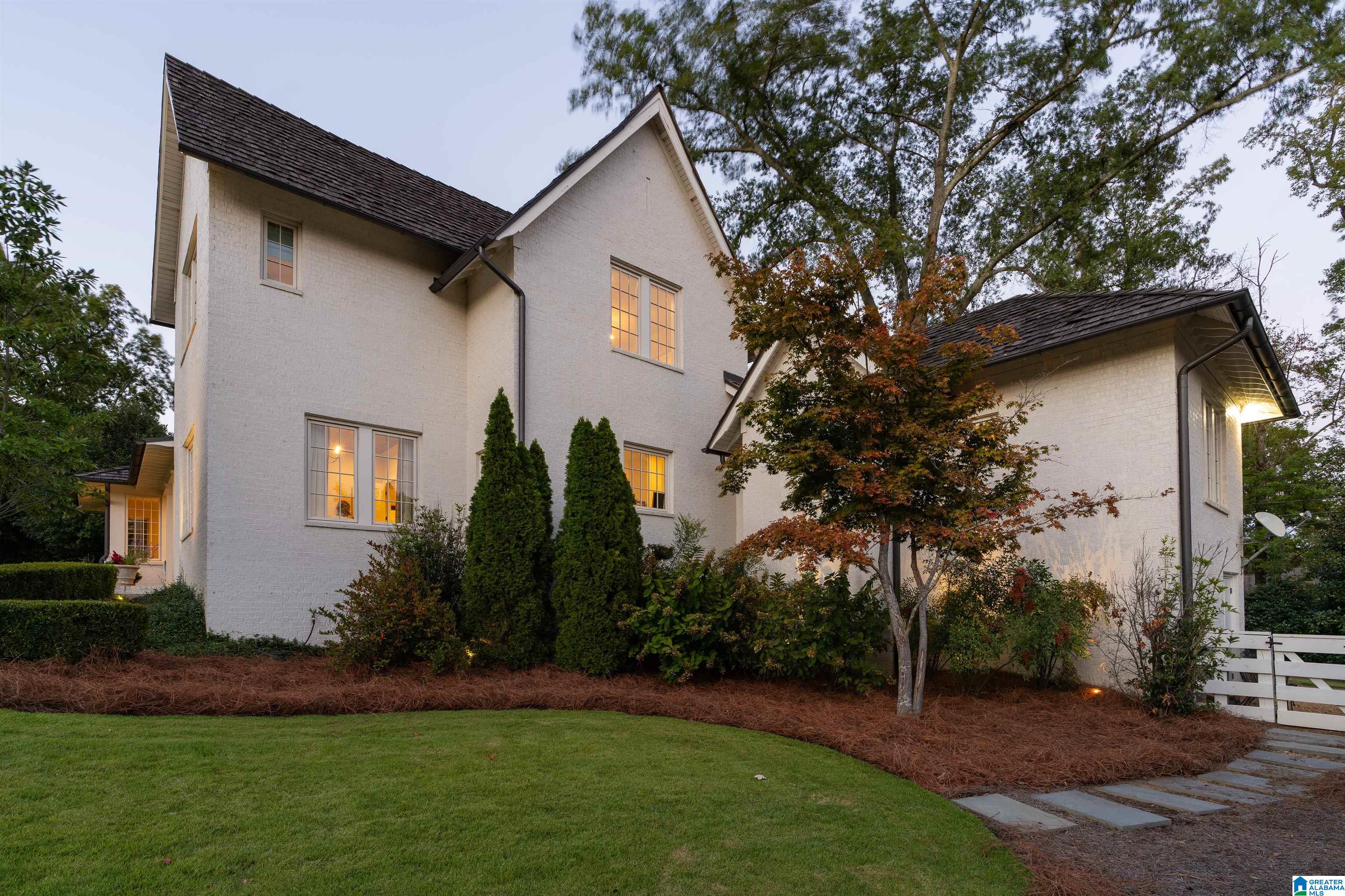 White brick two-story house with glowing windows, autumn trees, and a curved lawn at dusk.