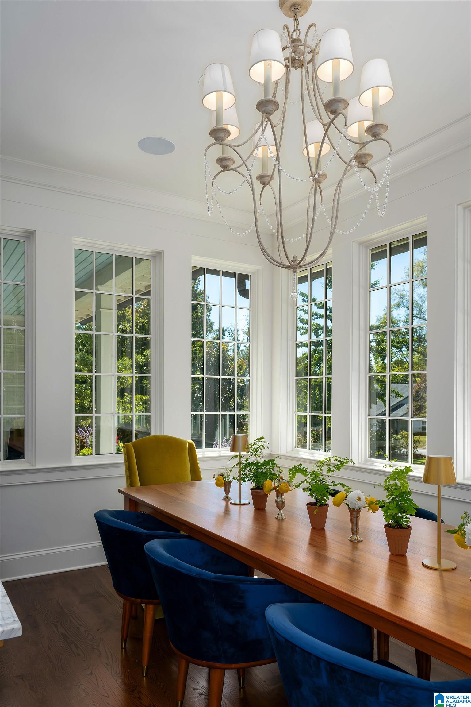 Dining room with tall windows, an ornate chandelier, and a wooden table surrounded by blue chairs.