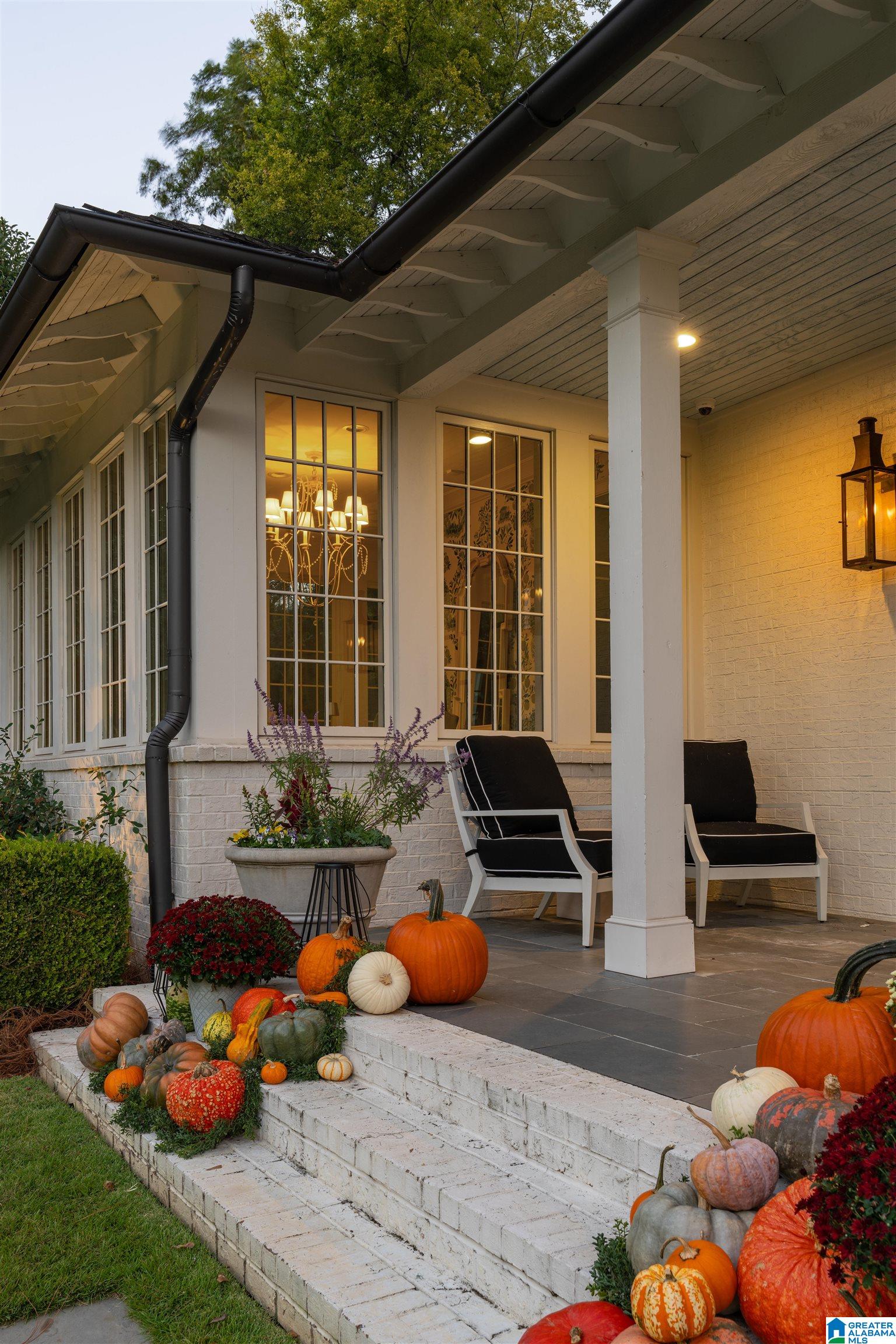 Covered white-brick porch with black chairs and autumn pumpkins lining the steps.