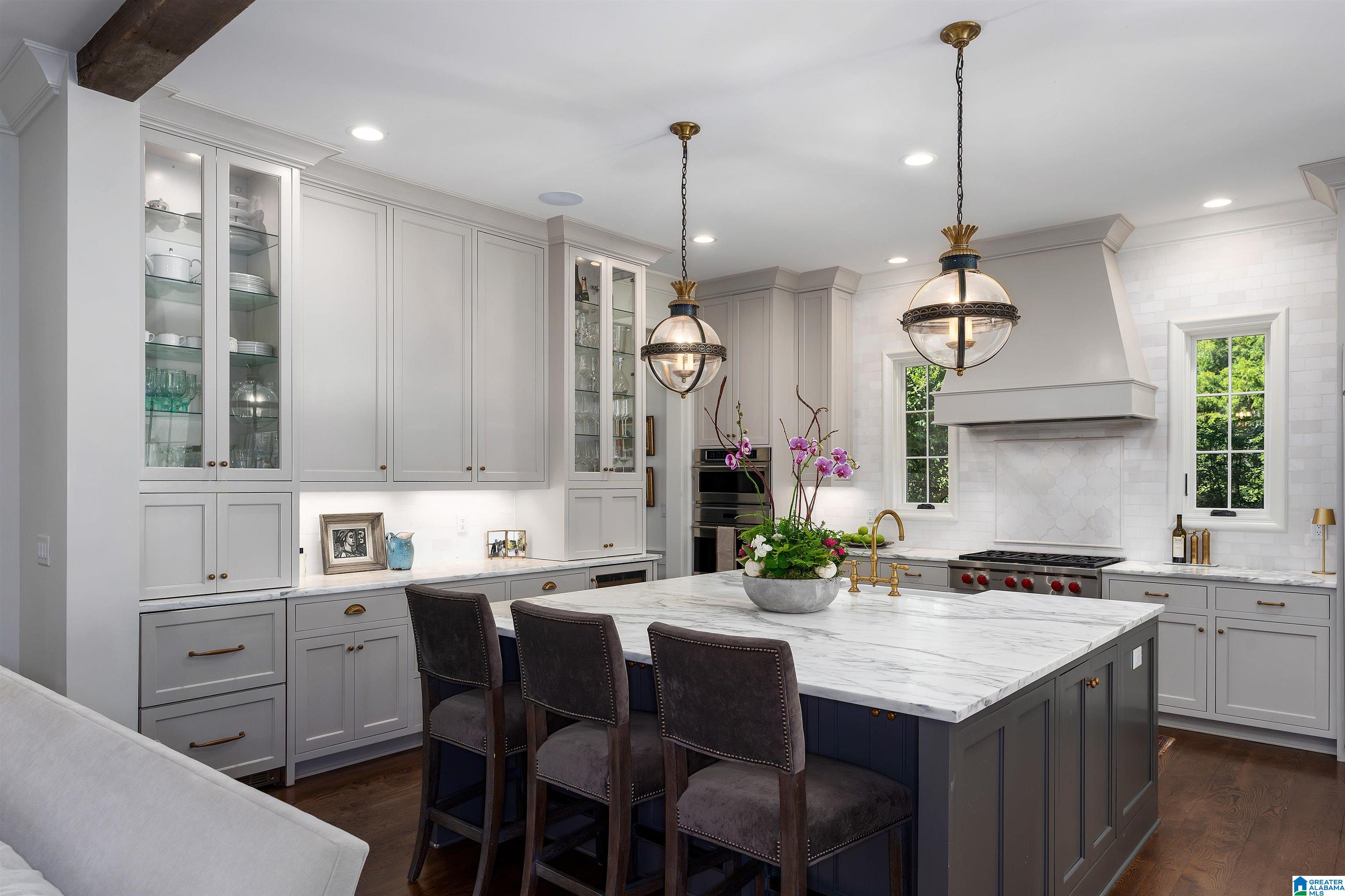Kitchen with a large marble island, gray cabinetry, brass pendants, and a white tiled range hood.