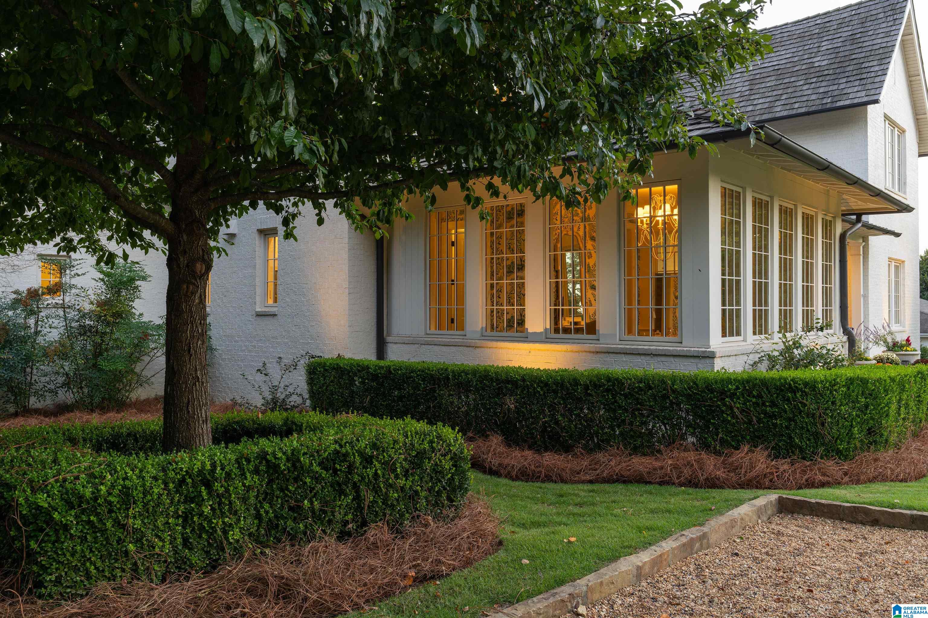 White-brick home exterior with tall illuminated windows, a mature shade tree, and clipped hedges.