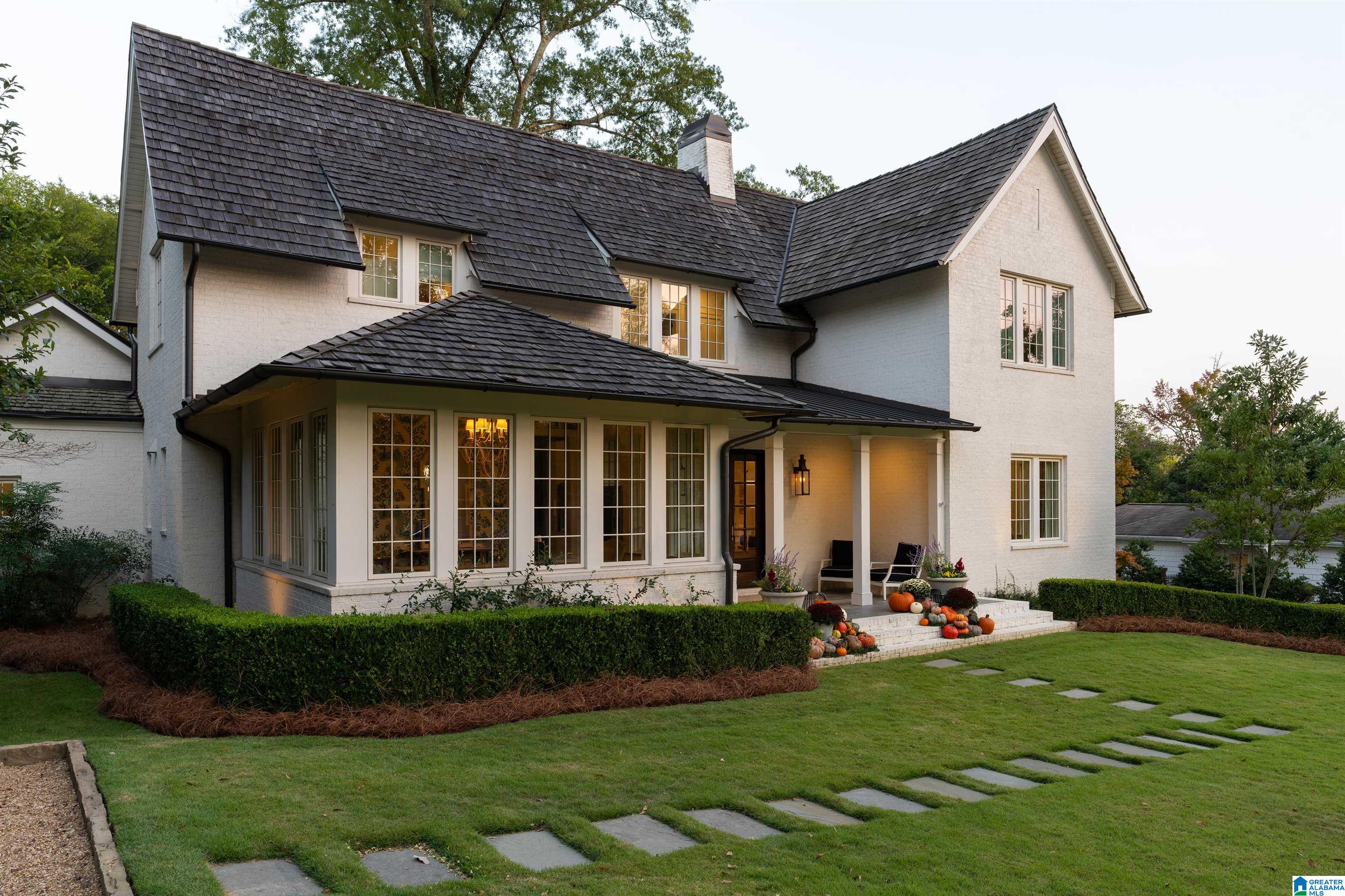 White brick two-story house with a dark roof, warmly lit windows, and pumpkins on the porch steps.